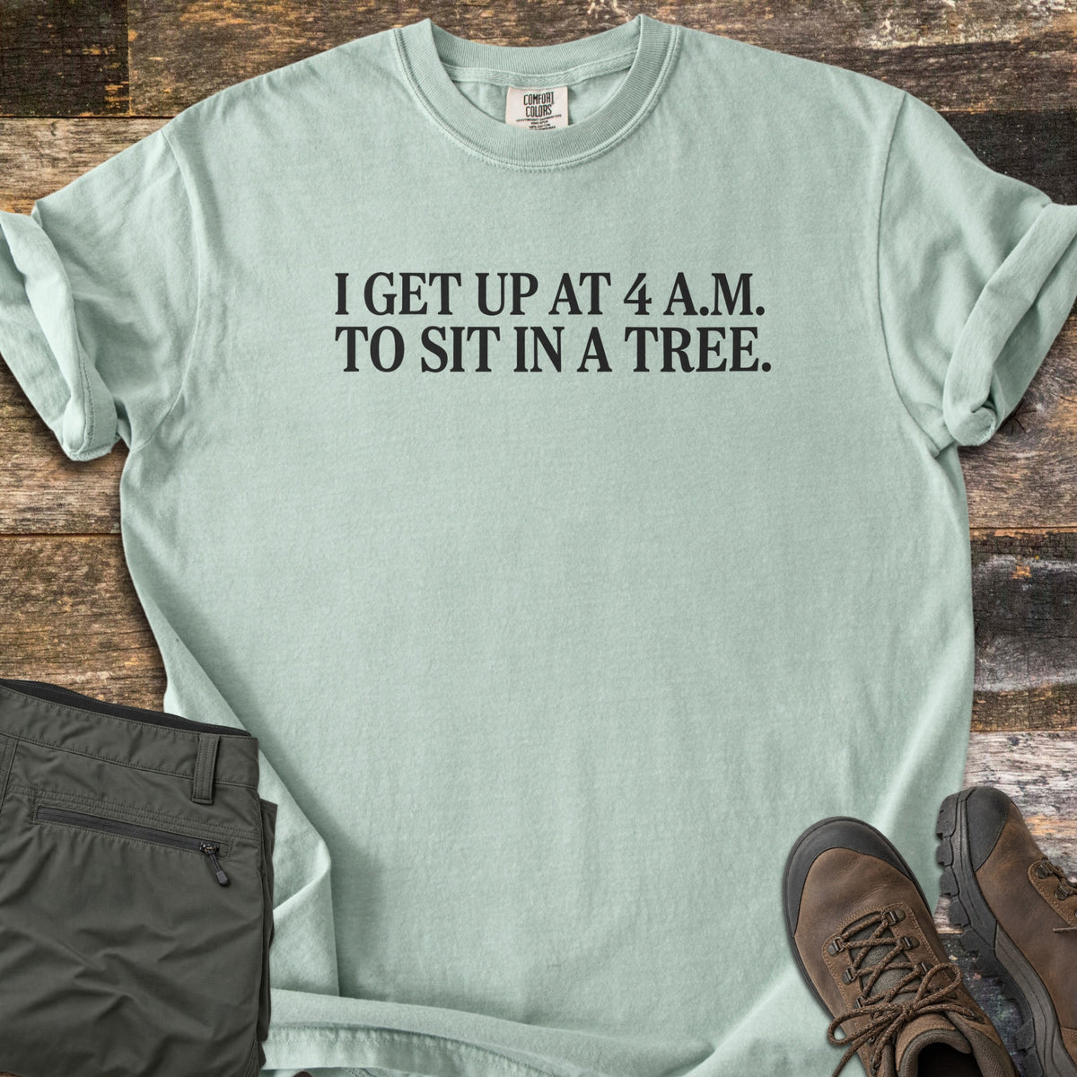 Sit In A Tree Comfort Colors T-shirt in Bay, laid flat. Black serif text reads I GET UP AT 4 A.M. TO SIT IN A TREE. across the chest. The shirt lies on weathered wood planks beside folded olive green cargo pants and brown leather hiking boots.