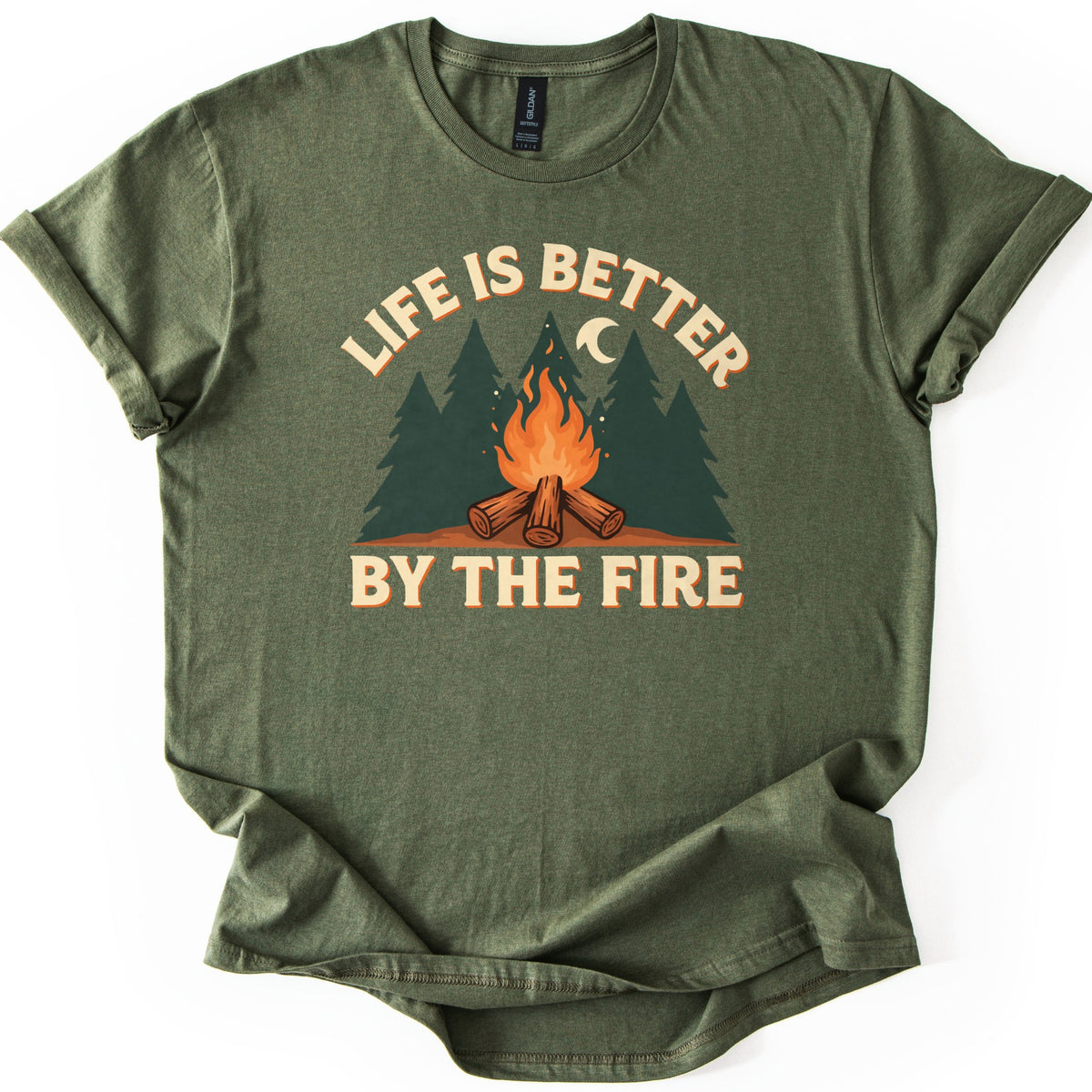 Life Is Better By The Fire Comfort Colors T-shirt in Military Green, laid flat. A campfire of crossed logs burns in front of dark green pine trees under a crescent moon, with LIFE IS BETTER arched above and BY THE FIRE below in cream letters. Cuffed sleeves, white background.