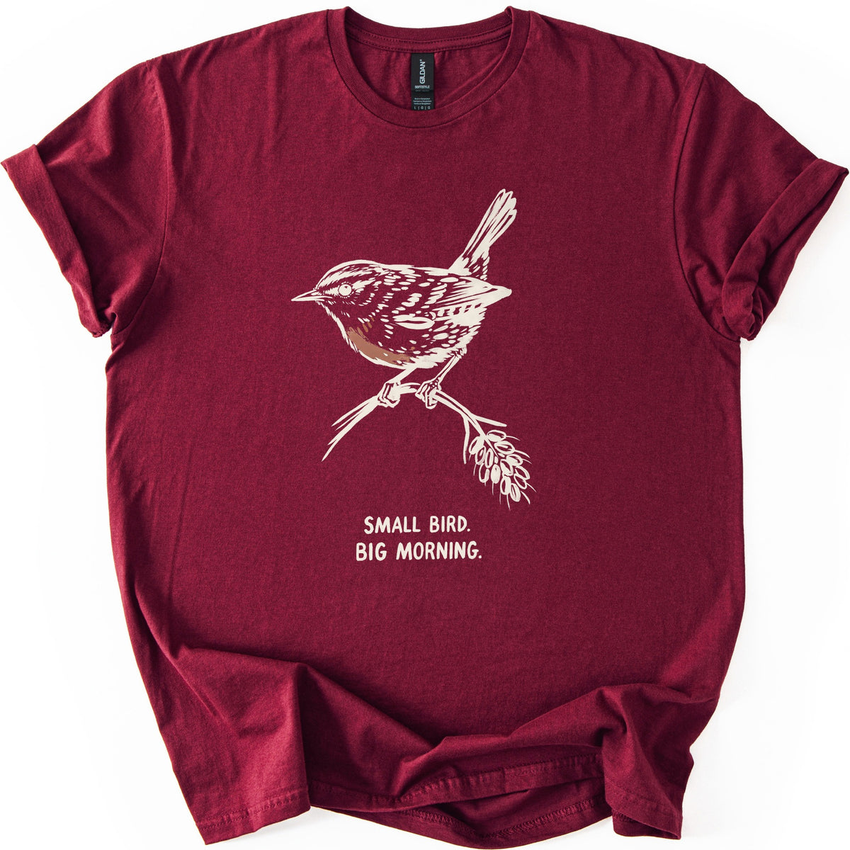 Small Bird Big Morning Comfort Colors T-shirt in Maroon, laid flat. A cream sketch of a wren with a brown-tinted breast perches on a twig with a seed head, above the words Small Bird. Big Morning. The sleeves are rolled up, on a white background.