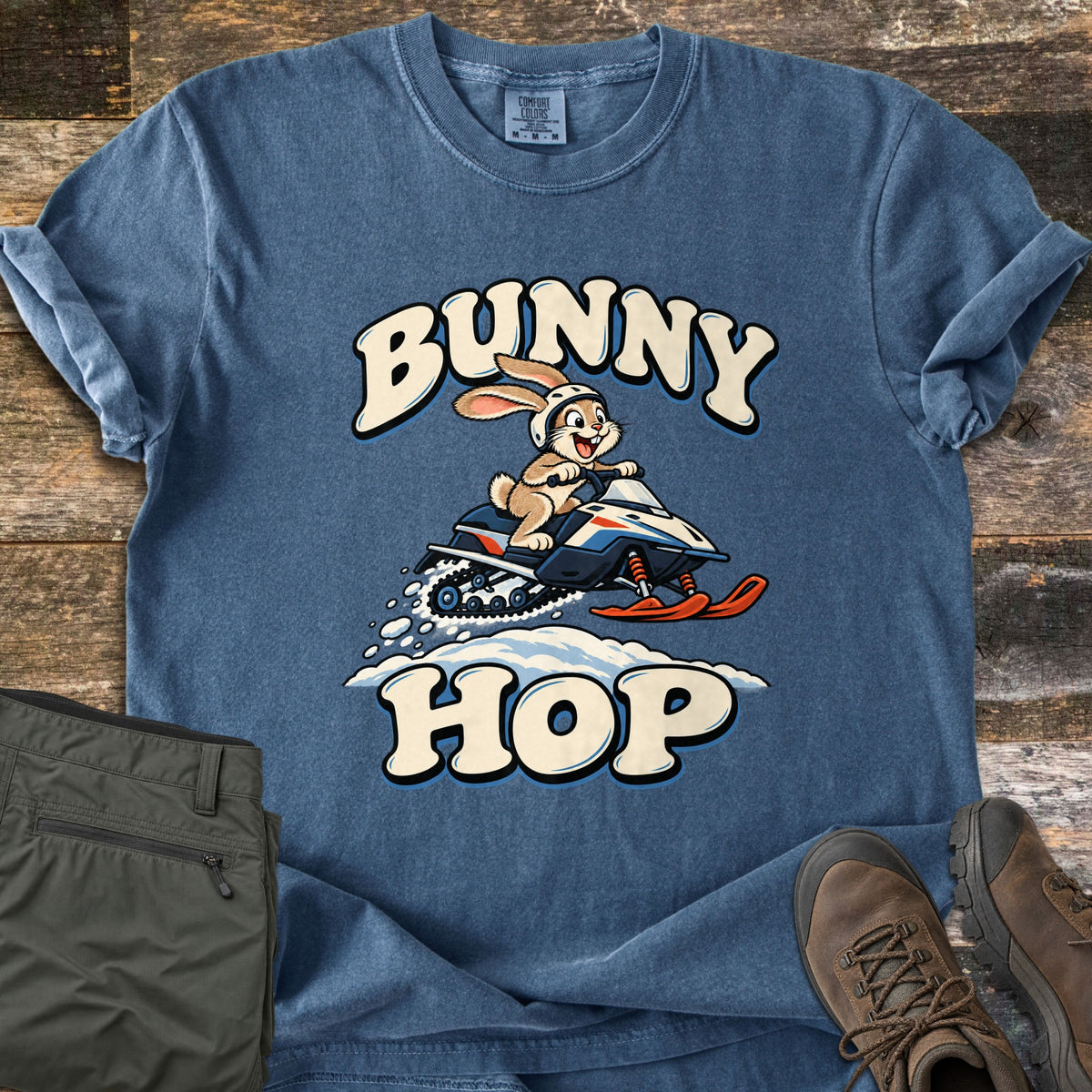 Bunny Hop Comfort Colors T-shirt in Denim, laid flat. A cartoon bunny in a white helmet rides a snowmobile, kicking up snow, between the words Bunny Hop. It lies on weathered wood beside olive pants and brown hiking boots.