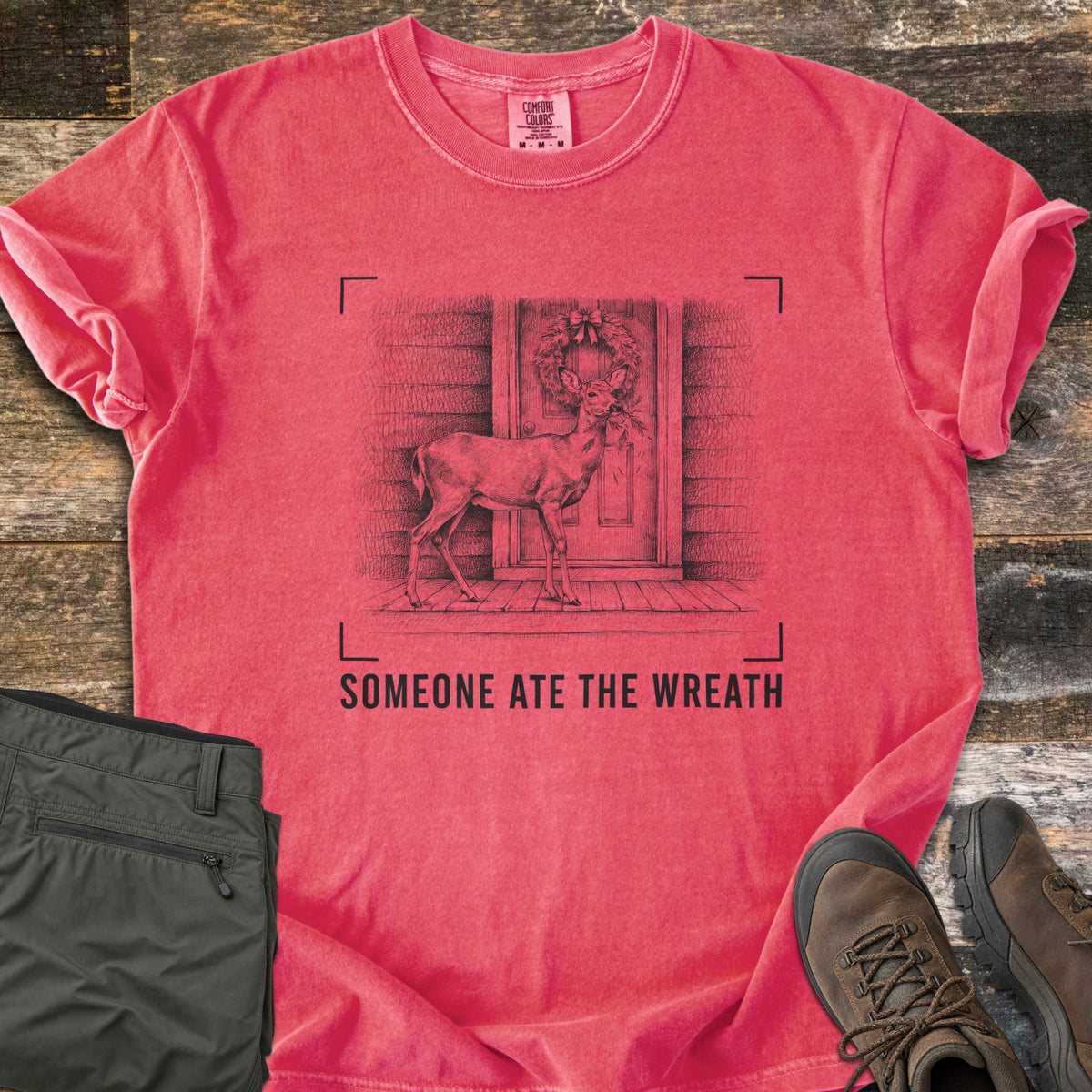 Someone Ate The Wreath Comfort Colors T-shirt in Watermelon, laid flat. A sketched deer nibbles a wreath on a cabin's front door, framed by corner marks above the words Someone Ate The Wreath. Shown on rustic wood planks with folded olive pants and brown hiking boots.