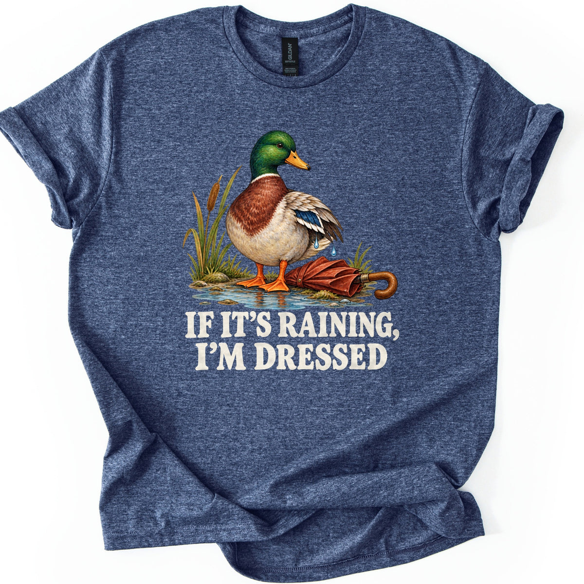 If It's Raining, I'm Dressed Comfort Colors T-shirt in Heather Navy, laid flat. A mallard duck with water dripping off its wing stands in a puddle by cattails beside a closed red umbrella, above the words IF IT'S RAINING, I'M DRESSED in cream. Cuffed sleeves, white background.