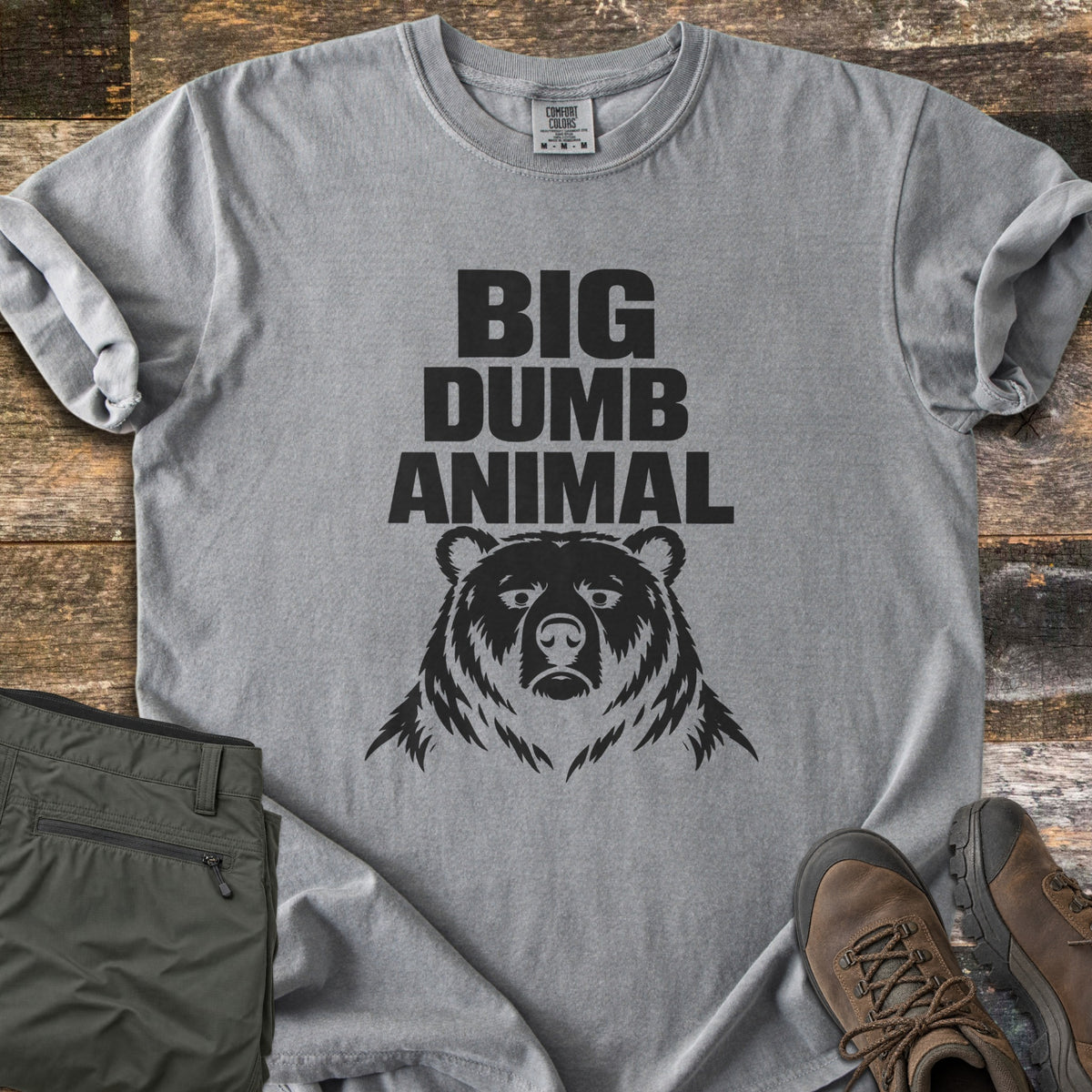 Big Dumb Animal Comfort Colors T-shirt in Grey, laid flat. Bold black block letters read Big Dumb Animal above a grumpy-faced bear head. The shirt, sleeves rolled, lies on weathered wood planks beside folded olive hiking pants and brown hiking boots.