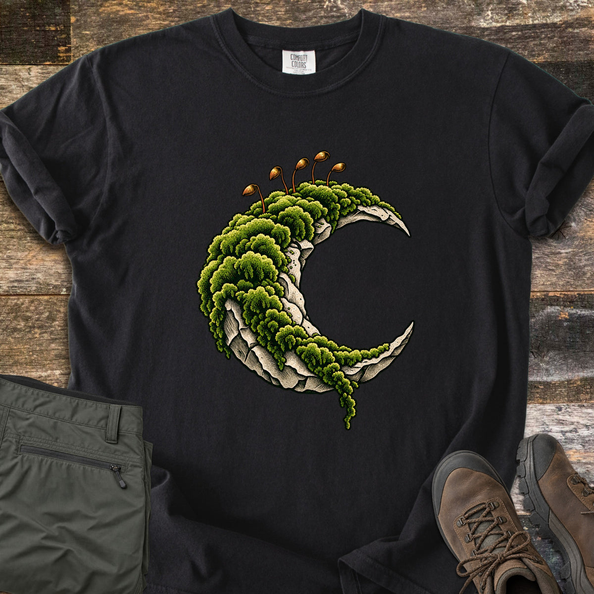 Moss Moon Comfort Colors T-shirt in Black, laid flat. A pale stone crescent moon covered in thick green moss, with thin brown spore stalks on top. Sleeves rolled, on dark weathered wood planks beside folded olive hiking pants and brown leather hiking boots.