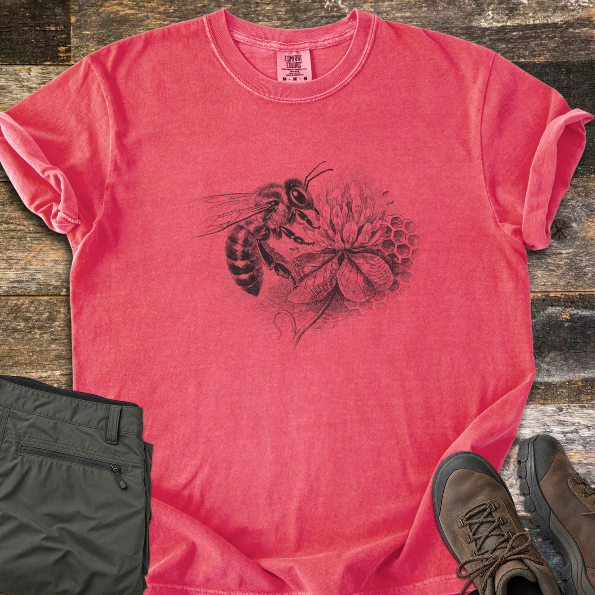 Honeybee On Clover Comfort Colors T-shirt in Watermelon, laid flat. A detailed black engraving-style illustration of a honeybee on a clover blossom with honeycomb cells behind it, no words. Sleeves rolled, on weathered wood planks beside olive hiking pants and brown hiking boots.