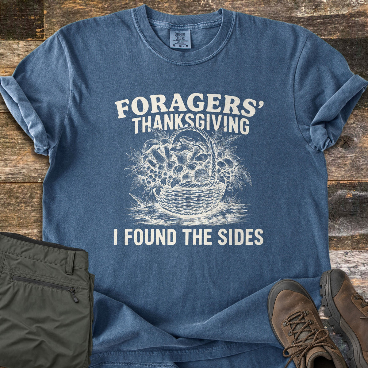 Foragers Thanksgiving Comfort Colors T-shirt in Denim, laid flat. A sketched basket of mushrooms, berries and leaves sits between the words Foragers' Thanksgiving and I Found The Sides. Olive hiking pants and brown hiking boots lie beside it on weathered wood planks.