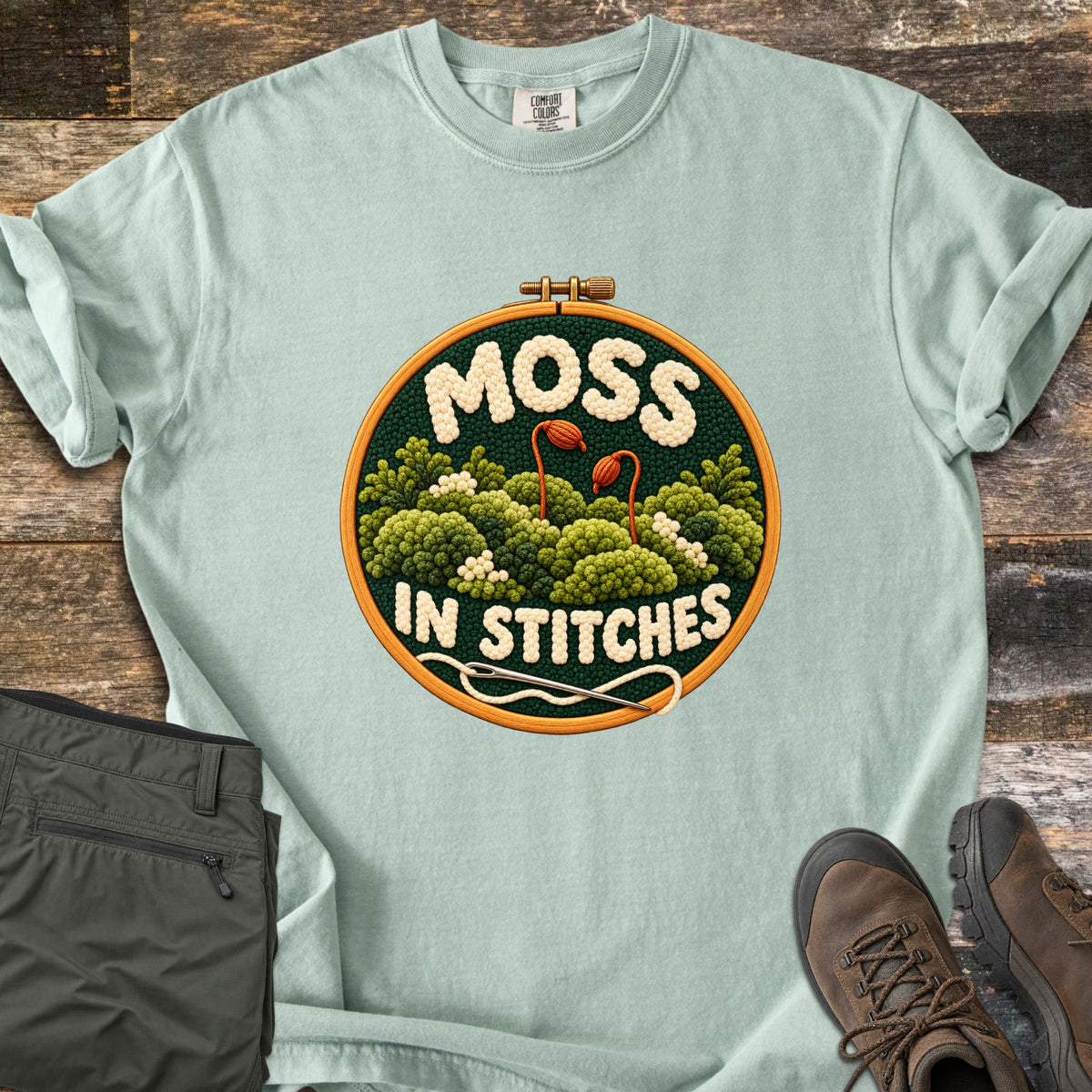 Moss in Stitches Comfort Colors T-shirt in Bay, laid flat. An embroidery hoop holds stitched green moss with two orange spore capsules, the word Moss above and In Stitches below, plus a needle and thread. Folded olive pants and brown hiking boots lie beside it on weathered wood.