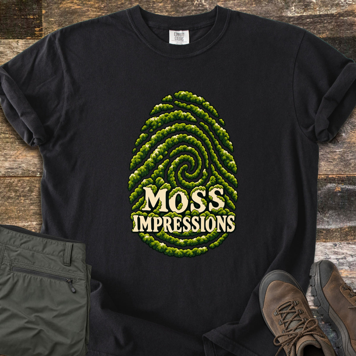 Moss Impressions Comfort Colors T-shirt in Black, laid flat. A fingerprint made of green moss ridges with the words Moss Impressions in cream letters. The shirt lies on weathered wood with folded olive hiking pants and brown hiking boots.