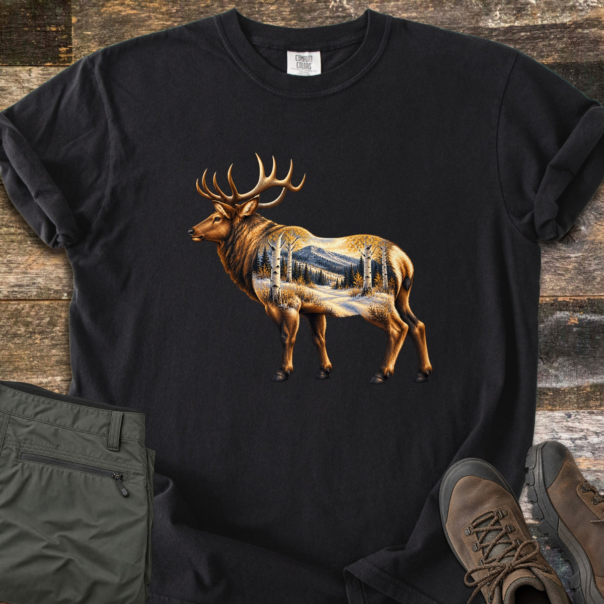 Aspen Pass Elk Comfort Colors T-shirt in Black, laid flat. A golden-brown bull elk in profile, its body filled with a snowy scene of aspen trees, pines and a mountain. Sleeves rolled, on rustic wood planks with folded olive hiking pants and brown hiking boots.