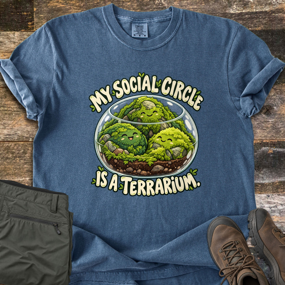 My Social Circle Is a Terrarium Comfort Colors T-shirt in Denim, laid flat. A glass terrarium holds three smiling moss balls on soil and stones, with the words My Social Circle Is a Terrarium. around it. Olive pants and brown hiking boots sit beside it on weathered wood.