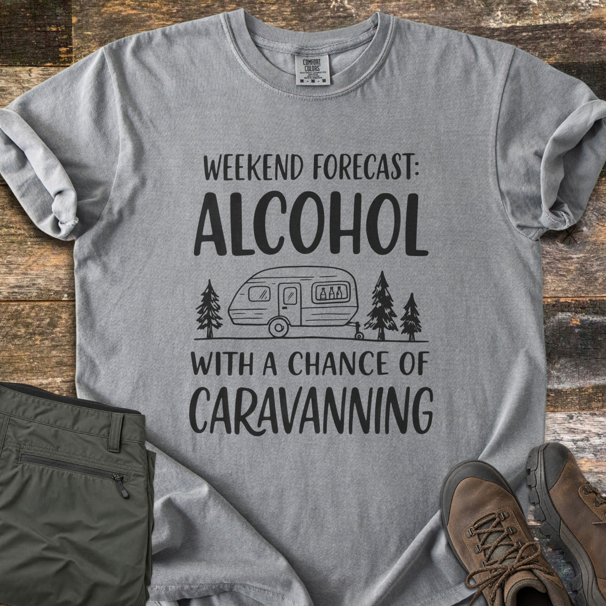 Alcohol With a Chance of Caravanning Comfort Colors T-shirt in Grey, laid flat. Black print reads Weekend Forecast: Alcohol above a caravan with bottles in its window between pine trees, then With a Chance of Caravanning. Olive trousers and brown hiking boots sit on dark wood planks.