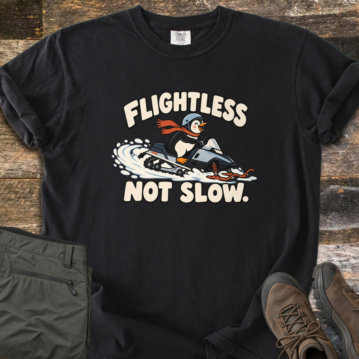 Flightless Not Slow Comfort Colors T-shirt in Black, laid flat. A cartoon penguin in a helmet and red scarf rides a snowmobile through snow between the words Flightless Not Slow. Folded olive pants and brown hiking boots sit beside it on weathered wood.