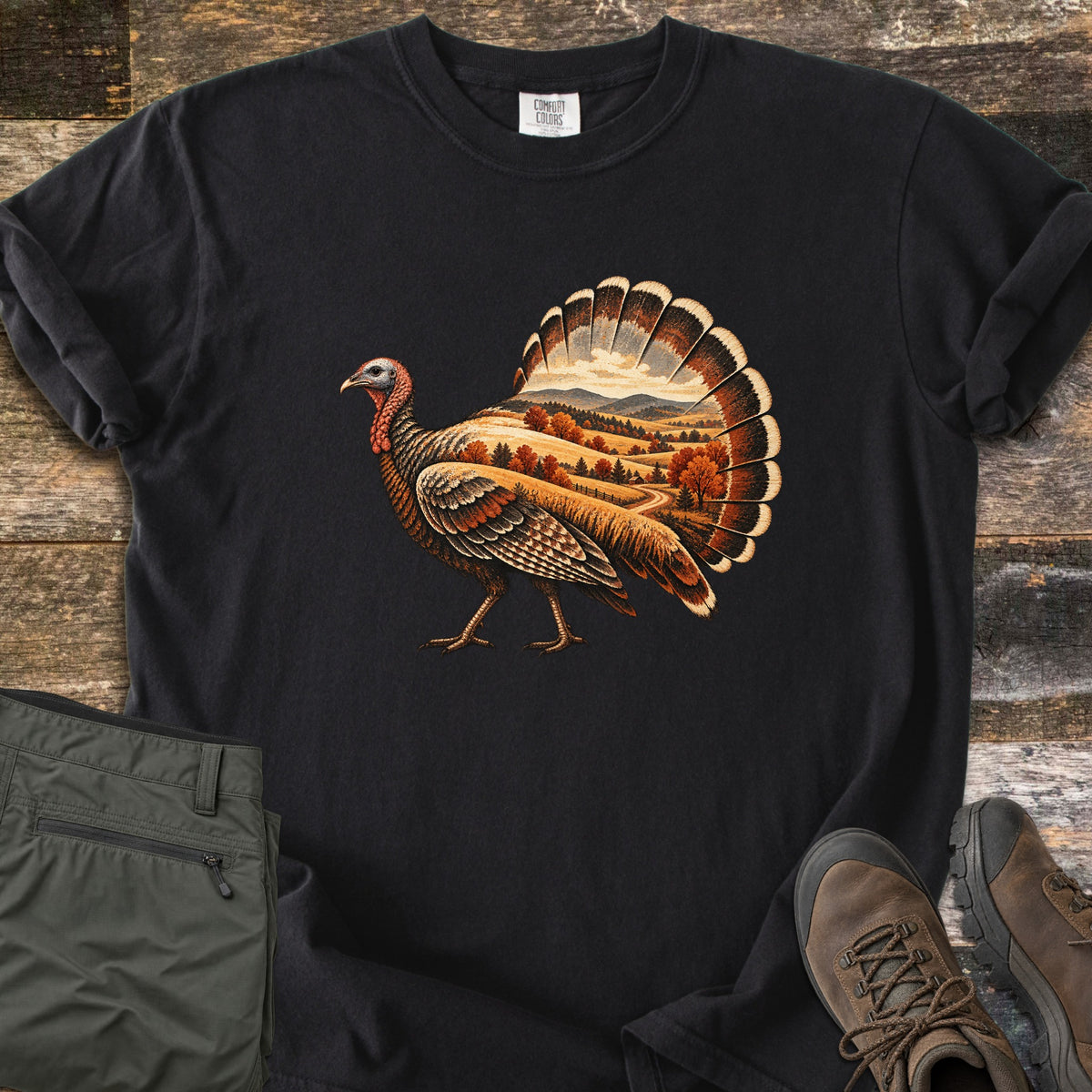 Turkey Harvest Hills Comfort Colors T-shirt in Black, laid flat. A walking wild turkey with an autumn landscape of golden hills, orange trees and a winding road inside its body and fanned tail. Sleeves rolled, on weathered wood with olive pants and brown hiking boots.