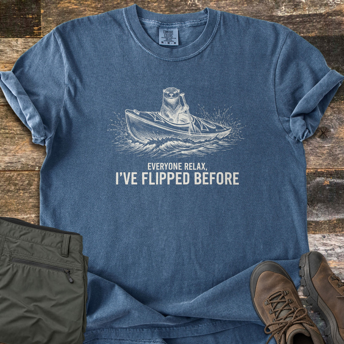 Everyone Relax I've Flipped Before Comfort Colors T-shirt in Denim, laid flat. A sketched otter paddles a kayak through splashing waves above the words Everyone Relax, I've Flipped Before. It lies on weathered wood with sleeves rolled, beside olive hiking pants and brown hiking boots.