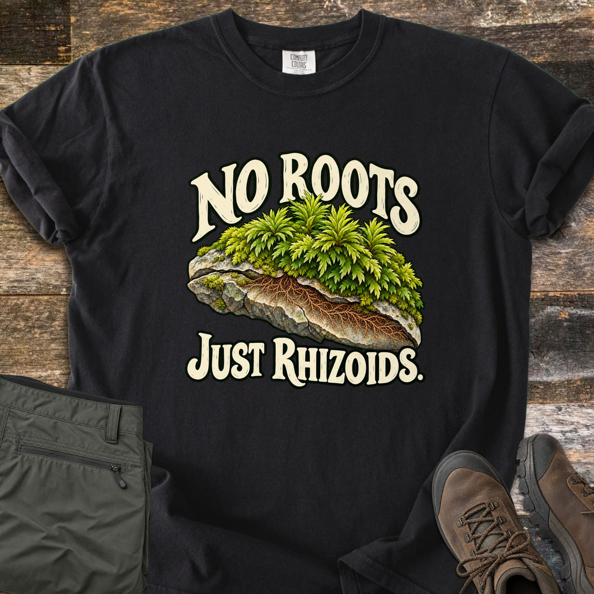 No Roots Just Rhizoids Comfort Colors T-shirt in Black, laid flat. Green moss sprouts from a cracked rock with brown rhizoids threading underneath, between the words No Roots Just Rhizoids. On rustic wood beside folded olive hiking pants and brown hiking boots.