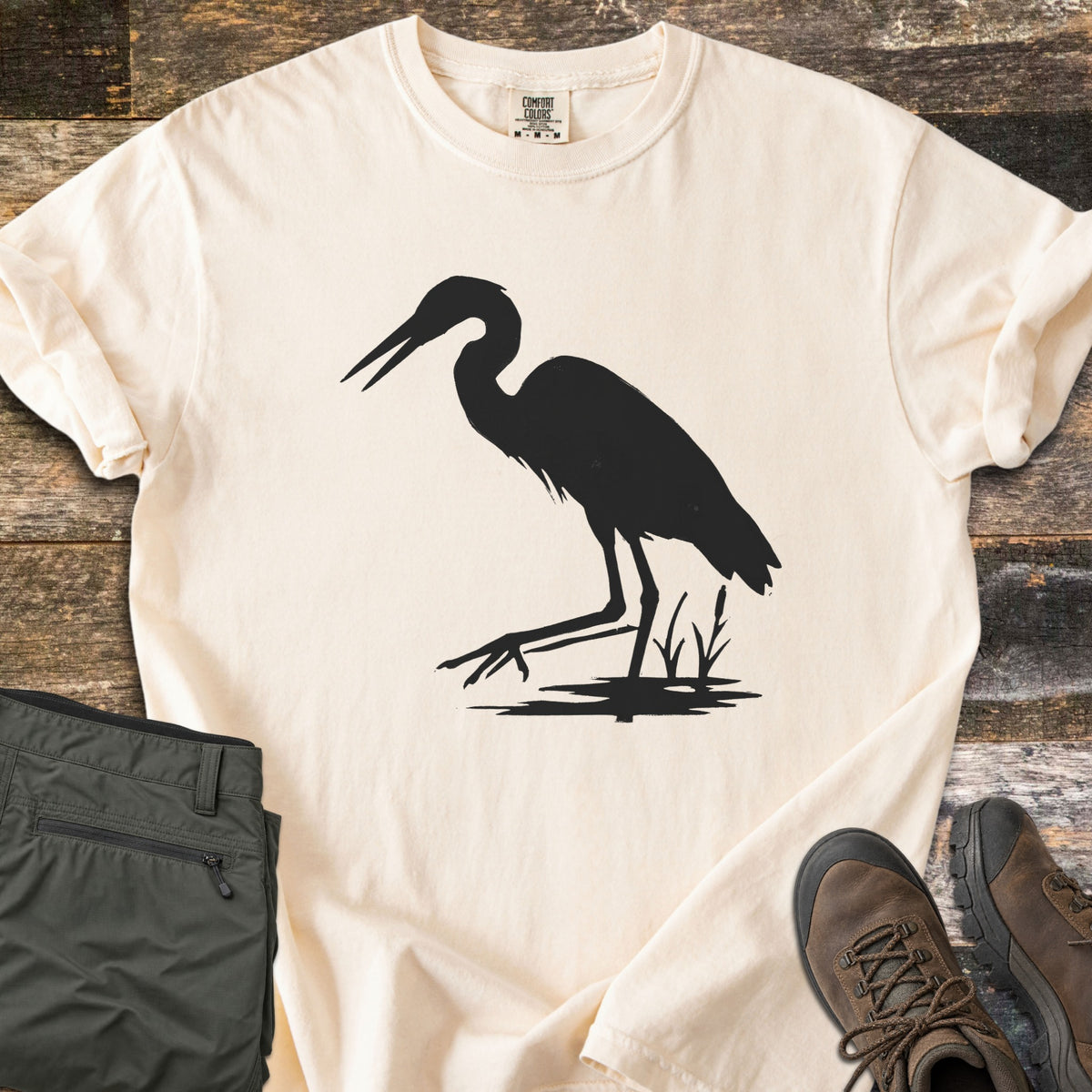 Great Blue Heron Comfort Colors T-shirt in Ivory, laid flat. A black silhouette of a heron wading with its beak open, one foot raised, beside cattails over a patch of water. Sleeves rolled, on weathered wood with olive hiking pants and brown hiking boots.