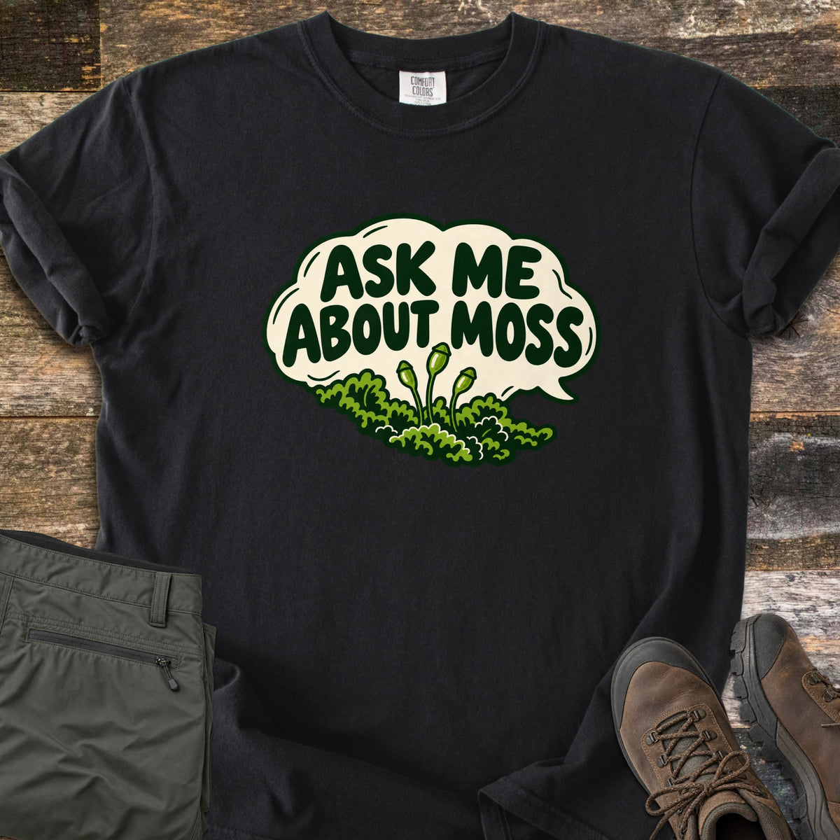 Ask Me About Moss Comfort Colors T-shirt in Black, laid flat. A cream speech bubble reads Ask Me About Moss in dark green letters above a mound of green moss with three spore stalks. Sleeves rolled, on weathered wood with folded olive hiking pants and brown hiking boots.