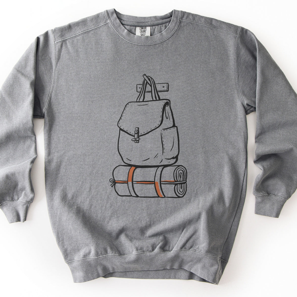 Backpack And Bedroll Comfort Colors crewneck sweatshirt in Grey, laid flat. Black line art of a canvas backpack with a toggle clasp hanging by its straps from a wall peg above a rolled bedroll tied with orange straps. No words are printed. Shown on a white background.