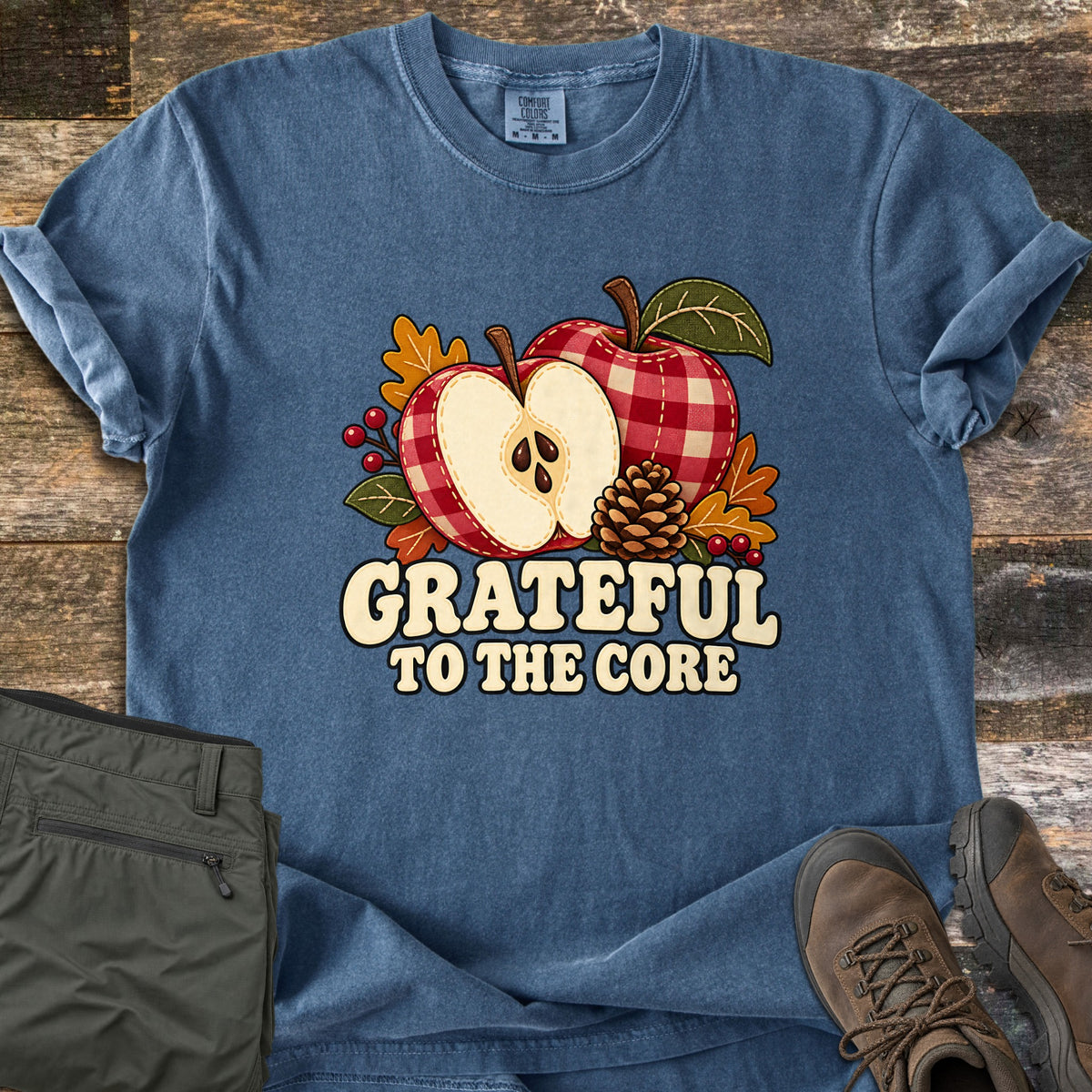 Grateful to the Core Comfort Colors T-shirt in Denim, laid flat. Two red gingham apples, one cut in half, with a pinecone, berries and autumn leaves above the words Grateful to the Core. Olive cargo pants and brown hiking boots rest beside it on weathered wood.
