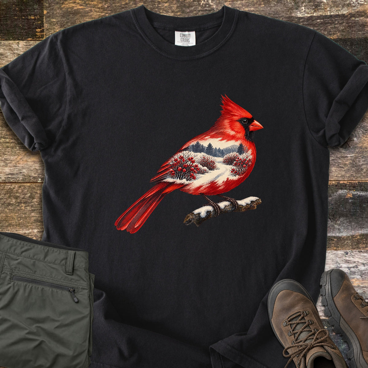 Winter Cardinal Comfort Colors T-shirt in Black, laid flat. A red cardinal perched on a snowy branch, its body filled with a snowy landscape of pine trees and red berry bushes. Rolled sleeves on weathered wood, with olive pants and brown hiking boots beside it.