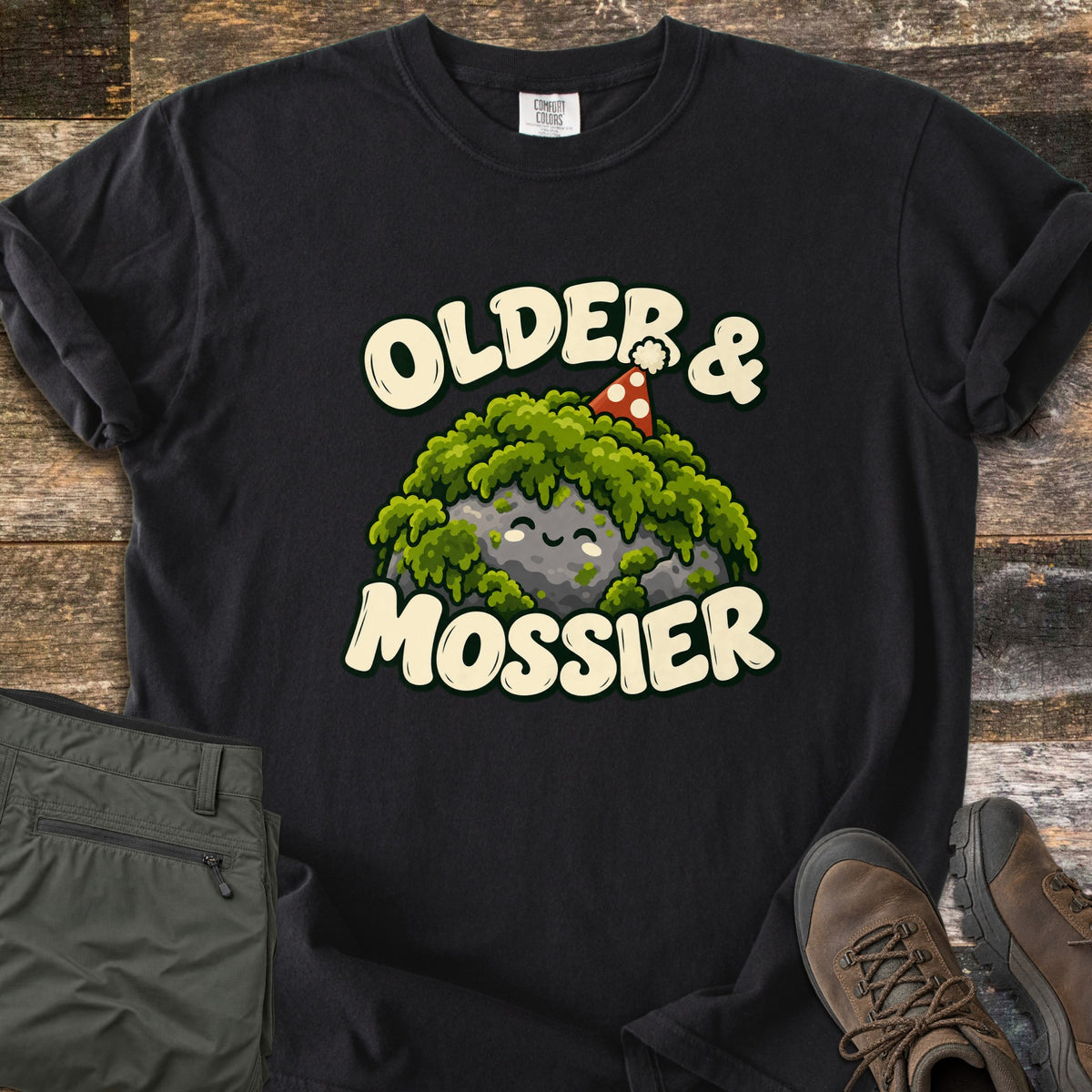 Older & Mossier Comfort Colors T-shirt in Black, laid flat. A smiling gray rock topped with green moss and a red polka-dot party hat sits between the words Older & Mossier in cream letters. Folded olive hiking pants and brown hiking boots rest on weathered wood planks.