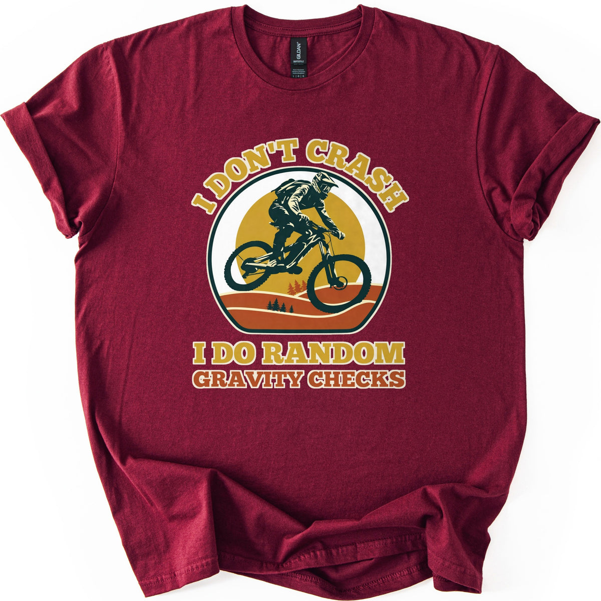Random Gravity Checks MTB Comfort Colors T-shirt in Maroon, laid flat. A mountain biker in a full-face helmet jumps against a mustard sun over orange hills with pine trees, framed by the words I Don't Crash above and I Do Random Gravity Checks below. Sleeves rolled, on white.