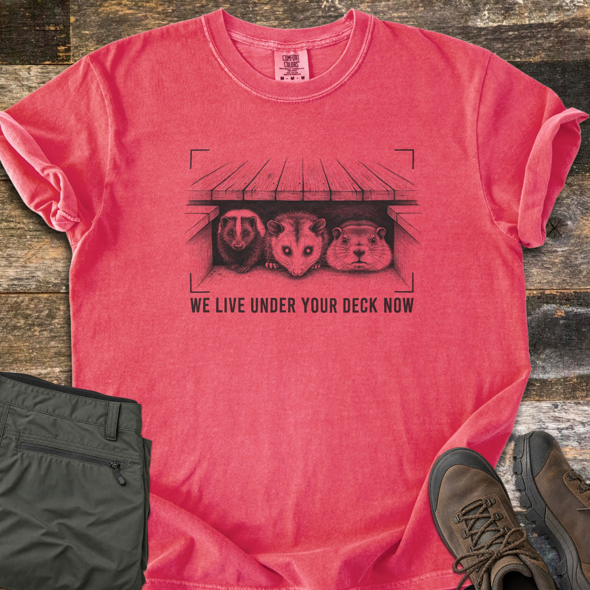 We Live Under Your Deck Now Comfort Colors T-shirt in Watermelon, laid flat. In black ink, a sketched skunk, opossum and groundhog peer from under a wooden deck inside viewfinder corners, above the words We Live Under Your Deck Now. Olive pants and brown hiking boots lie on weathered boards.