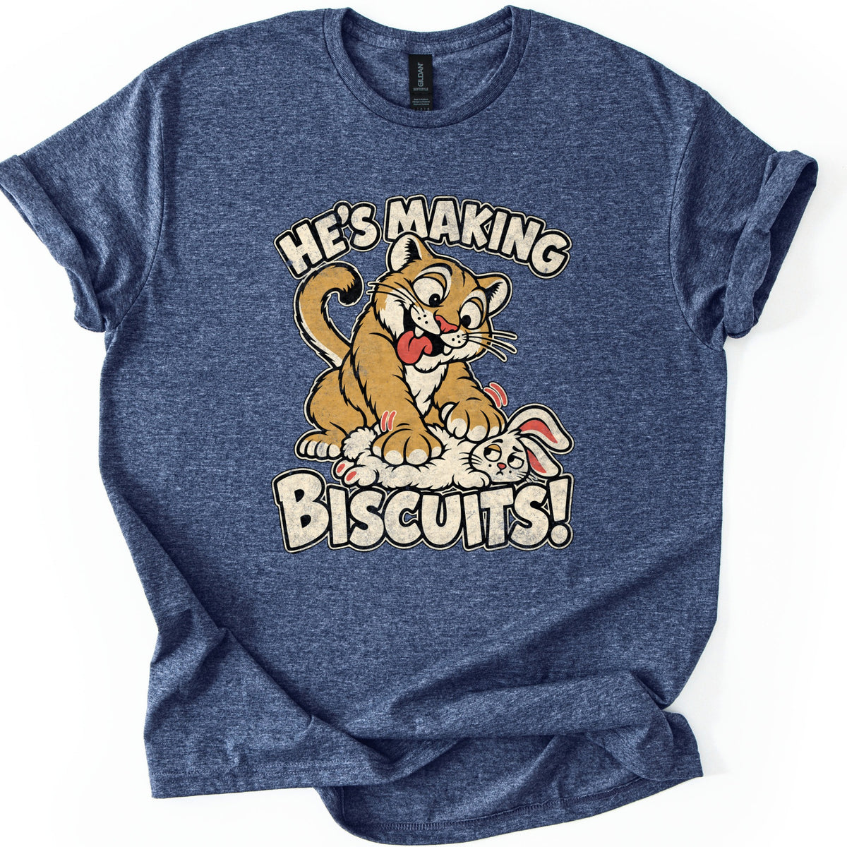 He's Making Biscuits Comfort Colors T-shirt in Heather Navy, laid flat. A grinning cartoon cougar with its tongue out kneads a dazed white rabbit on its back, with the words He's Making Biscuits! in distressed cream letters. The shirt lies on a white background with cuffed sleeves.