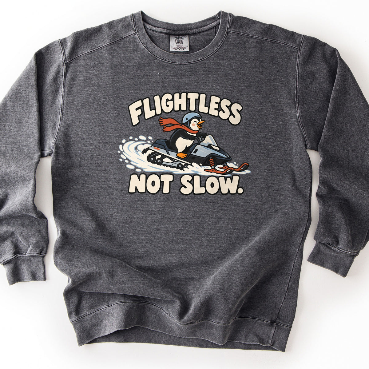 Flightless Not Slow Comfort Colors crewneck sweatshirt in Pepper, laid flat. A smiling cartoon penguin wearing a helmet and red scarf rides a snowmobile with orange skis through snow, with Flightless above and Not Slow. below in cream letters. It lies on a white background.