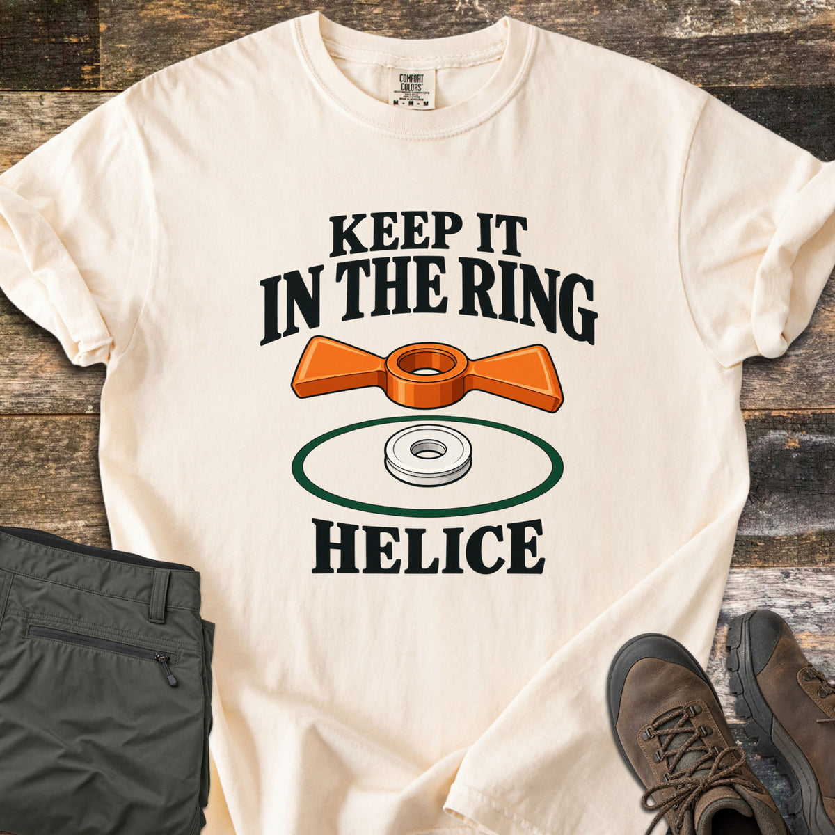Keep It in the Ring Comfort Colors T-shirt in Ivory, laid flat. An orange winged helice target floats above a white ring inside a green circle, between the words Keep It in the Ring and Helice. Olive pants and brown hiking boots sit beside it on weathered wood.