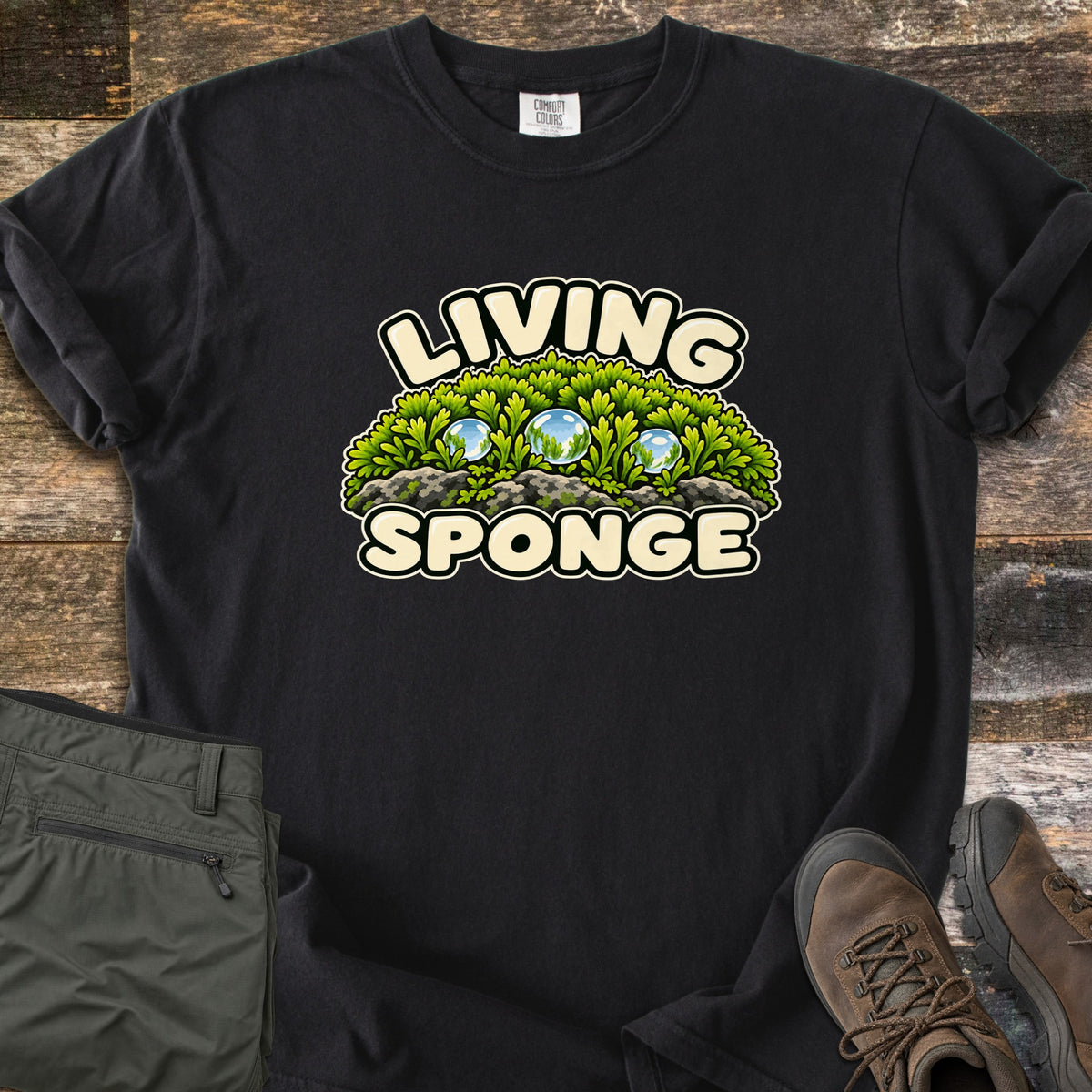 Living Sponge Comfort Colors T-shirt in Black, laid flat. A cartoon mound of bright green moss on gray rock, holding three clear water droplets, between the words Living Sponge. Olive hiking pants and brown leather hiking boots lie beside it on weathered wood.