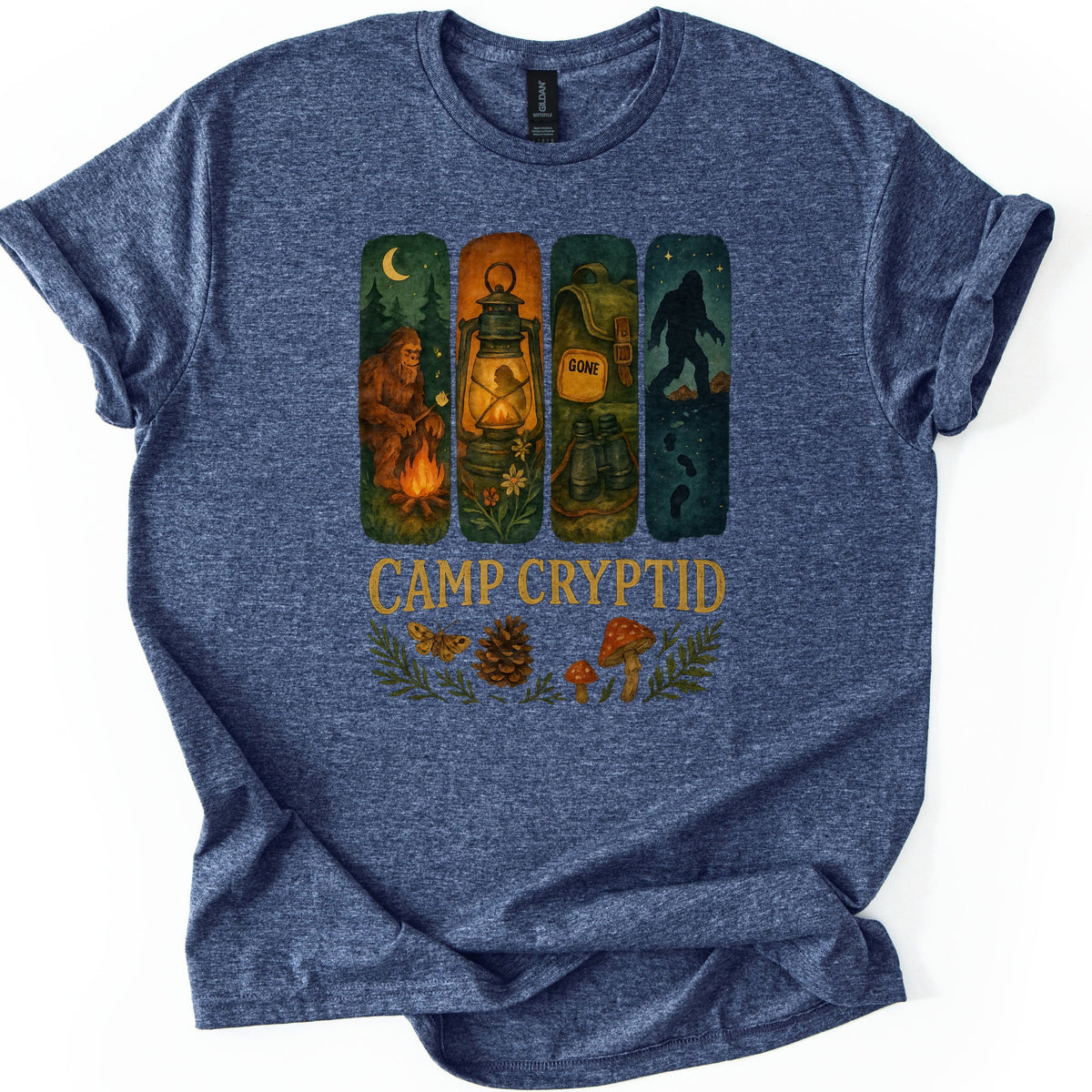 Camp Cryptid Big Foot Comfort Colors T-shirt in Heather Navy, laid flat. Four painted panels show Bigfoot at a campfire, a lit lantern, a backpack marked GONE, and Bigfoot's silhouette under stars, above the words Camp Cryptid and a moth, pinecone and mushrooms. Shown on a white background.