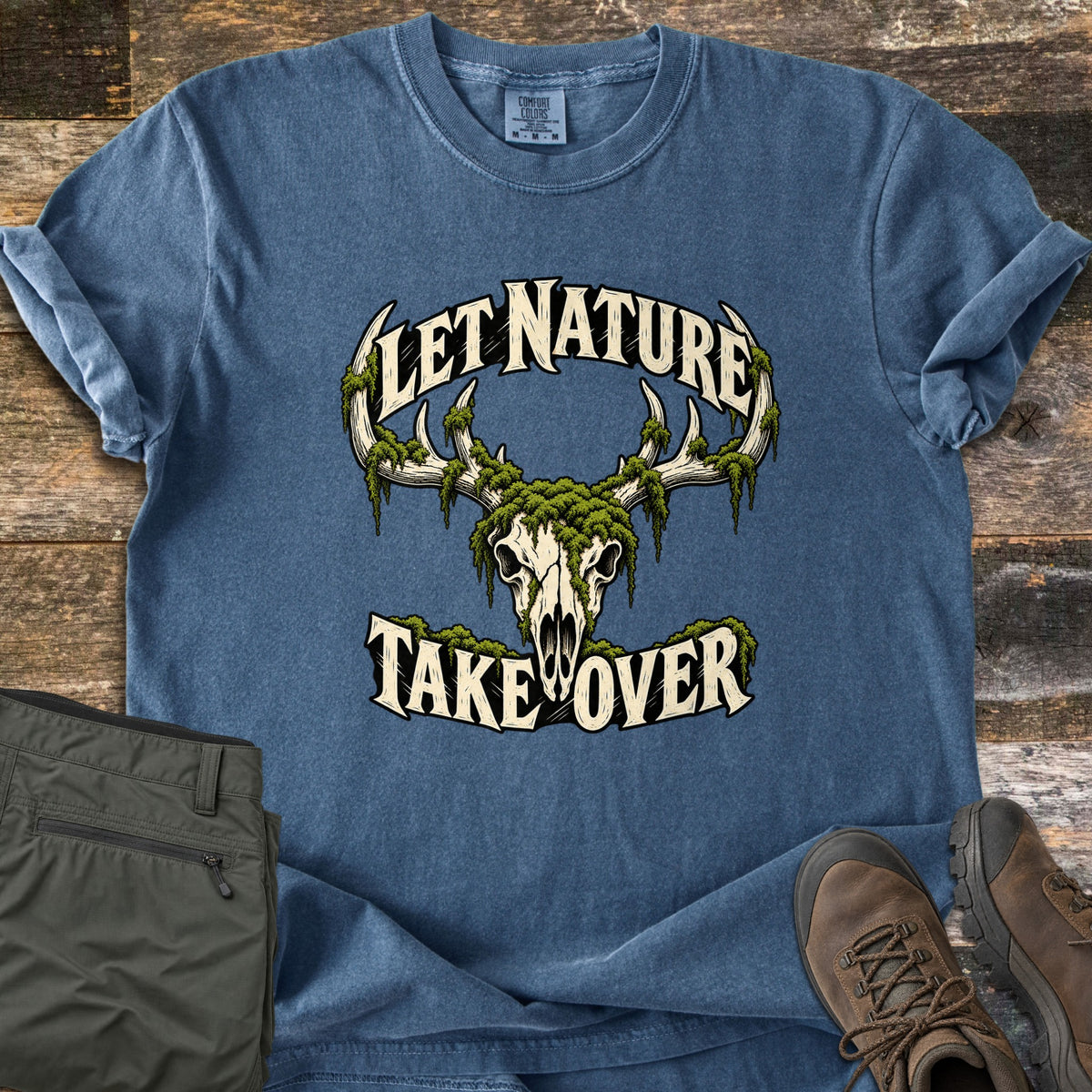 Let Nature Take Over Comfort Colors T-shirt in Denim, laid flat. A deer skull with antlers draped in green moss, with the words Let Nature arched above and Take Over below. Olive cargo pants and brown hiking boots lie beside it on weathered wood planks.