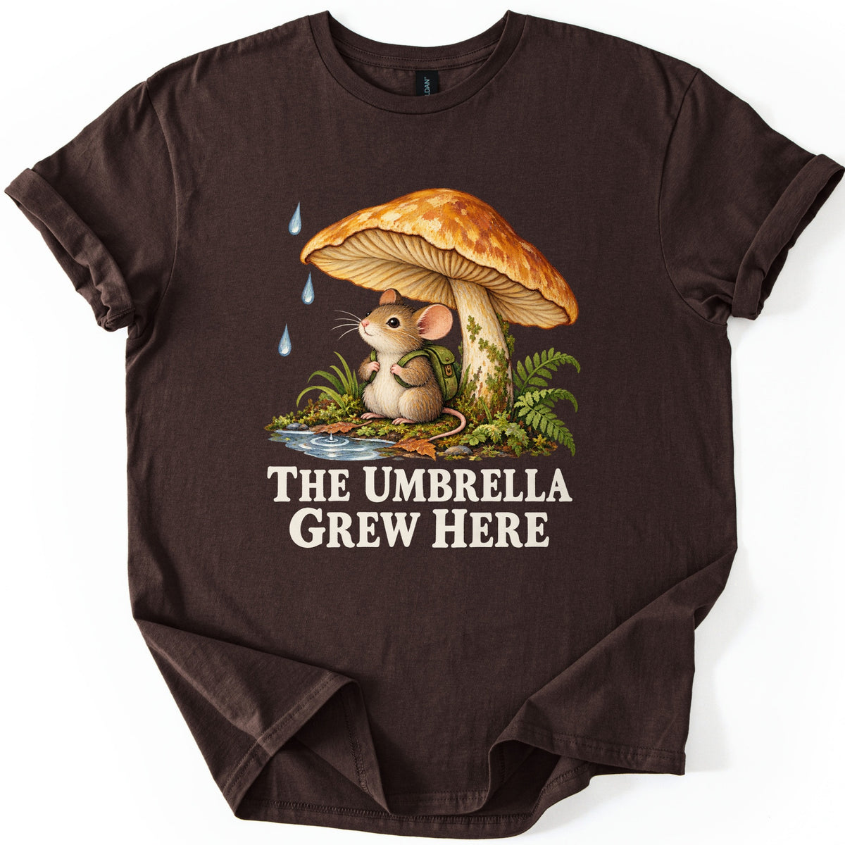 The Umbrella Grew Here Comfort Colors T-shirt in Dark Chocolate, laid flat. A mouse wearing a green backpack sits under a large orange mushroom as raindrops fall, beside ferns and a puddle, above the words The Umbrella Grew Here in cream letters. Shown on a white background.