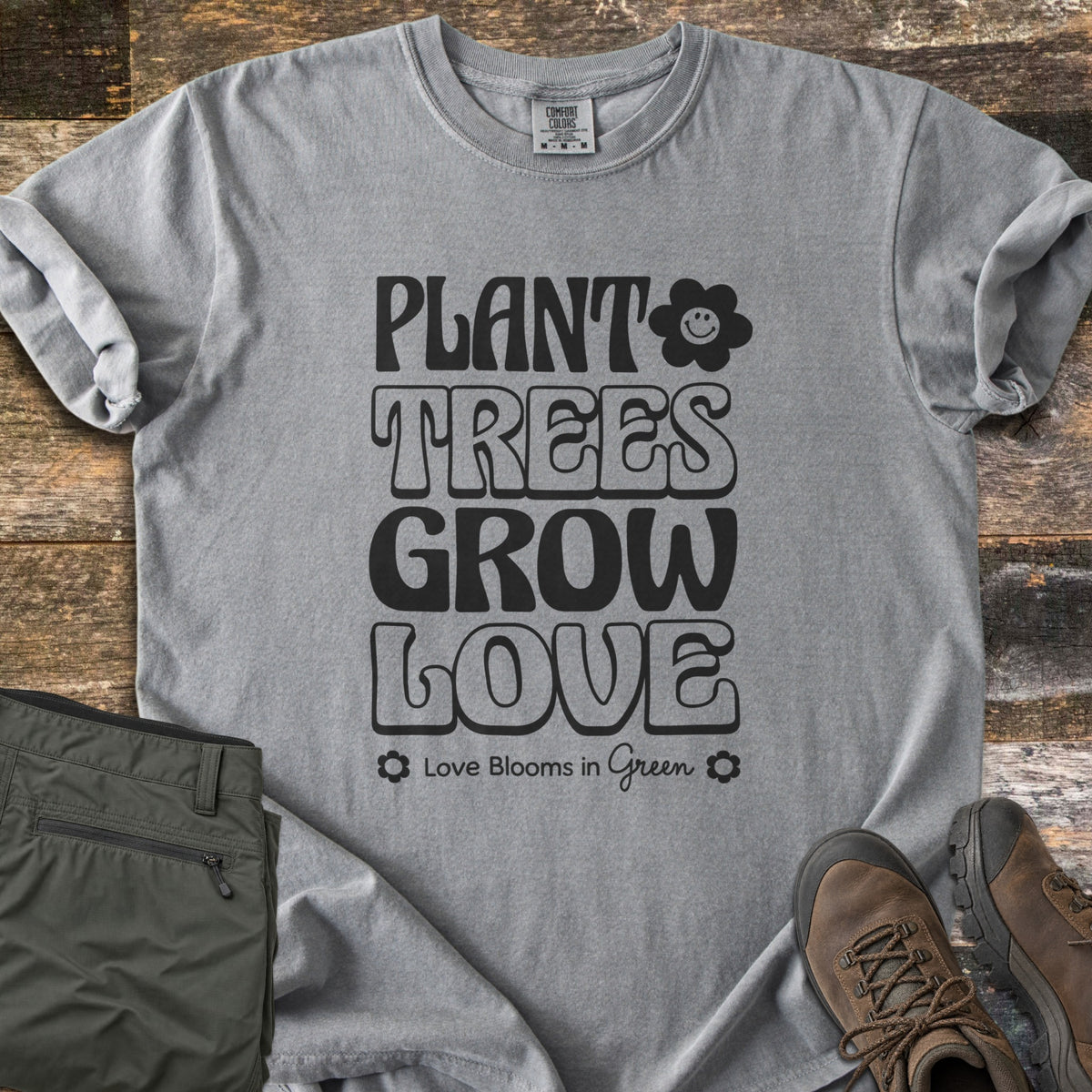 Plant Trees Grow Love Comfort Colors T-shirt in Grey, laid flat. Black retro bubble letters read Plant Trees Grow Love beside a smiling daisy, above Love Blooms in Green. Sleeves cuffed on weathered wood with folded olive hiking pants and brown hiking boots.