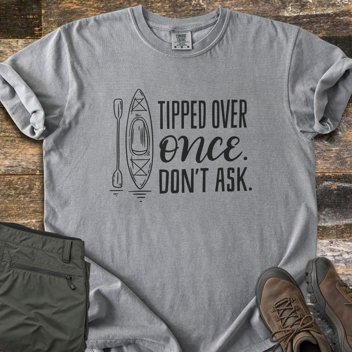 Tipped Over Once. Don't Ask. Comfort Colors T-shirt in Grey, laid flat. A black line drawing of a kayak and paddle sits beside the words Tipped Over Once. Don't Ask., with Once in script. Sleeves rolled, on weathered wood with folded olive hiking pants and brown hiking boots.