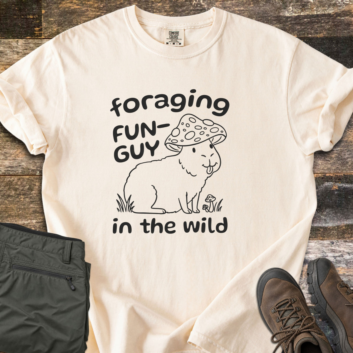Foraging Fun-Guy in the Wild Comfort Colors T-shirt in Ivory, laid flat. A black line drawing of a capybara wearing a spotted mushroom cap, tongue out, sits in grass by tiny mushrooms between the words foraging FUN-GUY in the wild. Olive hiking pants and brown boots lie on weathered wood.
