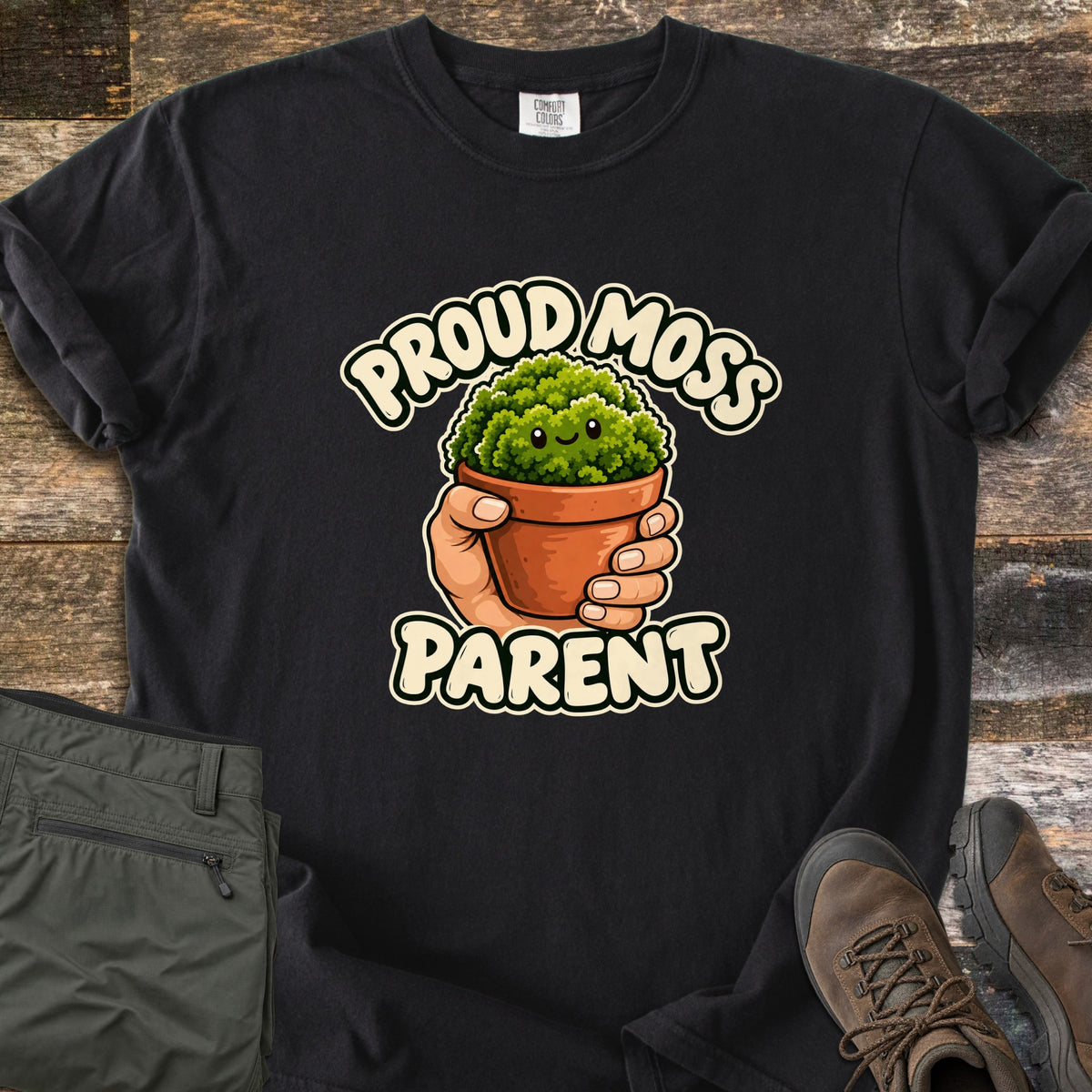 Proud Moss Parent Comfort Colors T-shirt in Black, laid flat. A cartoon hand holds a terracotta pot of smiling green moss between the words Proud Moss Parent. Sleeves are rolled, with green hiking pants and brown hiking boots on a weathered wood background.