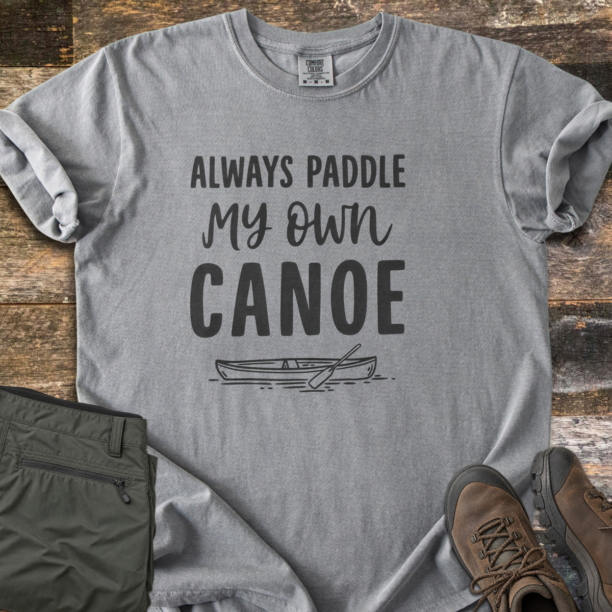 Paddle My Own Canoe Comfort Colors T-shirt in Grey, laid flat. Charcoal text reads Always Paddle My Own Canoe above a line drawing of a canoe with a paddle on the water. It lies on weathered wood with rolled sleeves, folded olive hiking pants, and brown hiking boots.