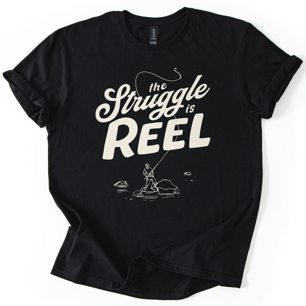 Struggle Is Reel Angler Comfort Colors T-shirt in Black, laid flat. Cream text reads the Struggle is REEL, with a fishing line arcing over the words to a hook. Below, a line drawing of an angler in a hat casts from a rock in the water. Sleeves cuffed, on a white background.