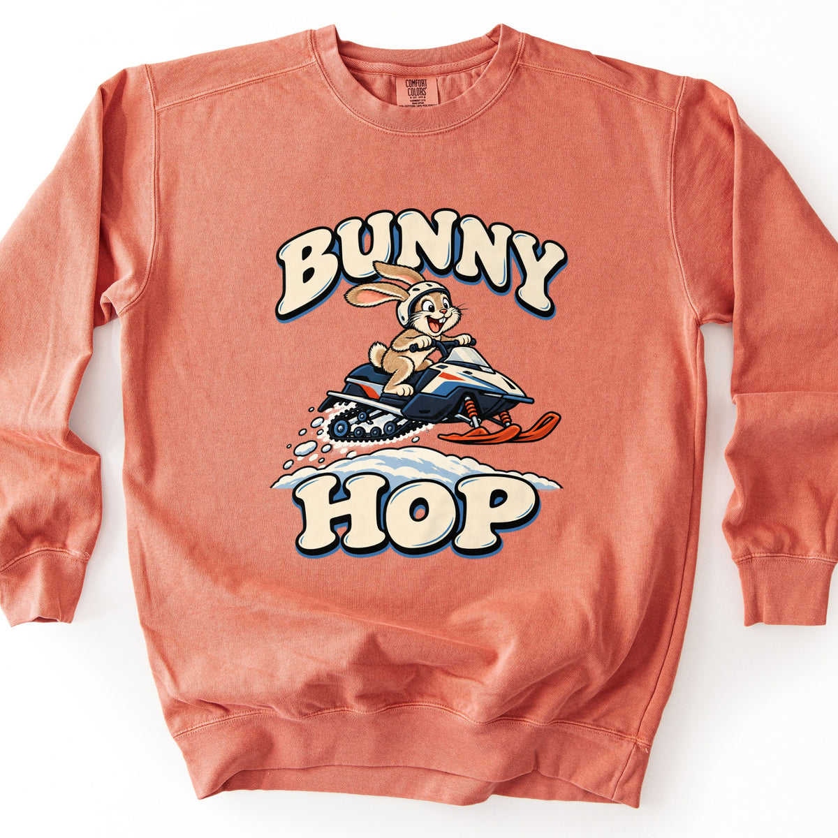 Bunny Hop Comfort Colors crewneck sweatshirt in Terracotta, laid flat. A grinning cartoon bunny in a white helmet rides a navy, white and orange snowmobile over a snowbank between the words Bunny Hop in cream bubble letters. The sweatshirt lies on a white surface.