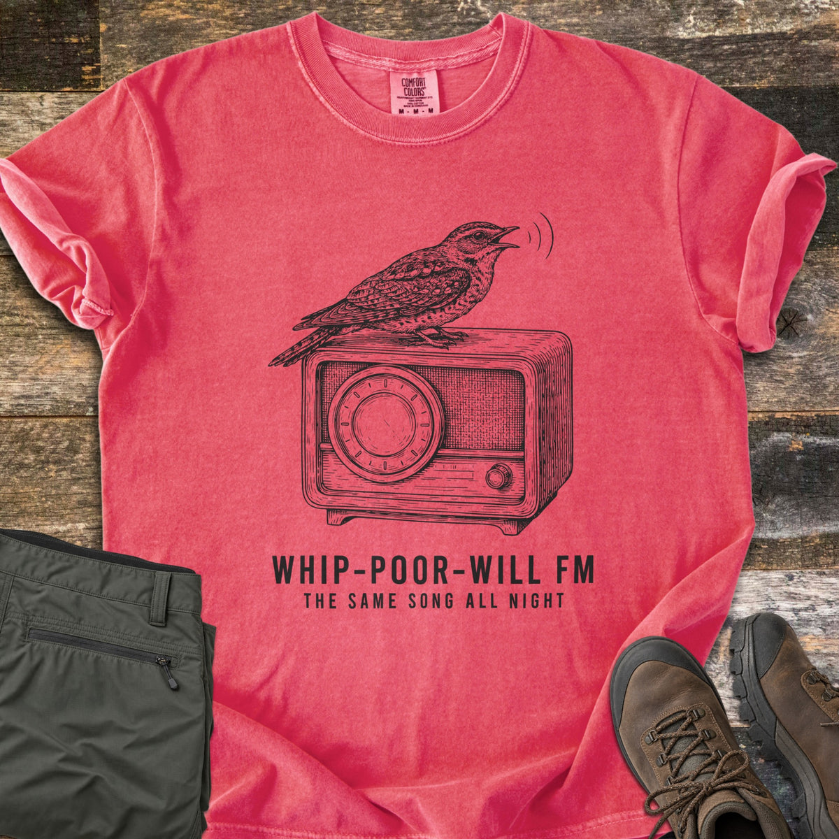 Whip-Poor-Will FM Comfort Colors T-shirt in Watermelon, laid flat. A sketched whip-poor-will sings atop a vintage radio above the words Whip-Poor-Will FM, The Same Song All Night. It lies with rolled sleeves on weathered wood beside folded olive pants and brown hiking boots.