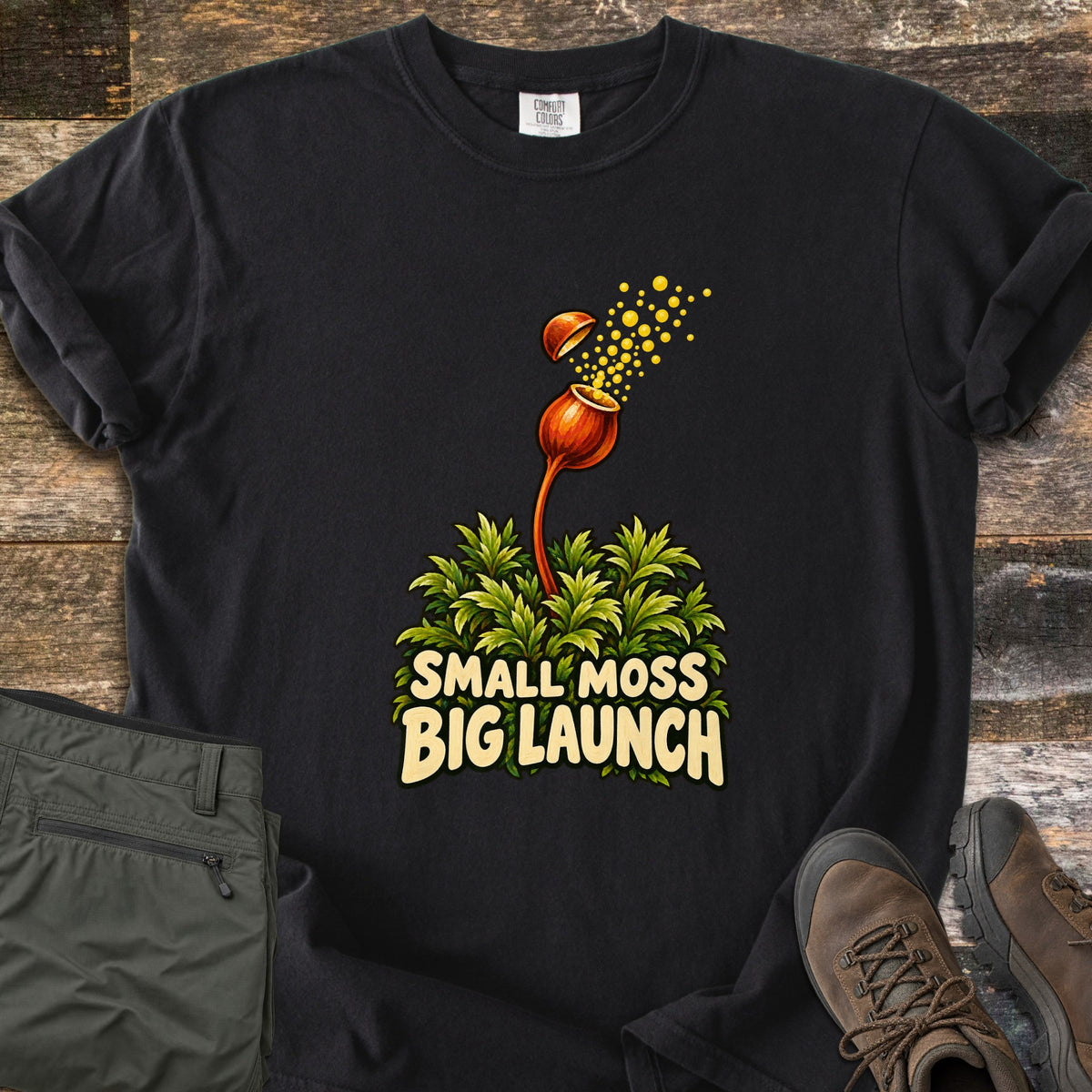 Small Moss Big Launch Comfort Colors T-shirt in Black, laid flat. A green moss clump sends up an orange capsule that pops its lid and bursts yellow spores above the words Small Moss Big Launch. The sleeves are rolled, with olive hiking pants and brown boots on rustic wood.