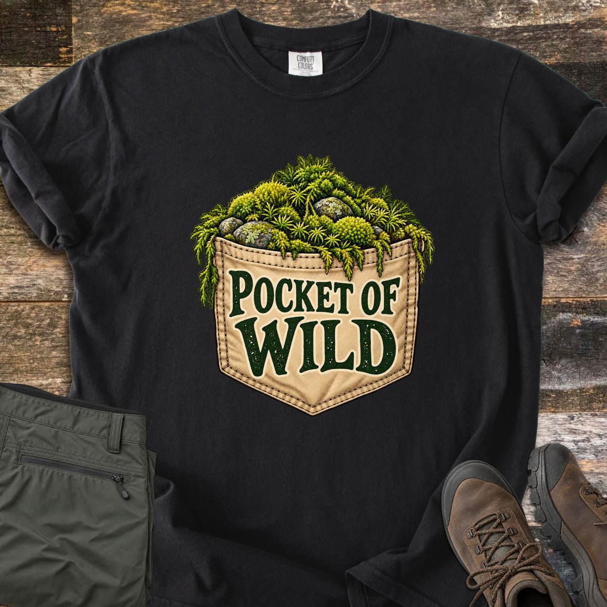 Pocket of Wild Comfort Colors T-shirt in Black, laid flat. A tan stitched jeans pocket overflowing with green moss and lichen-covered stones, printed with the words Pocket of Wild. Shown on weathered wood with folded olive hiking pants and brown hiking boots.