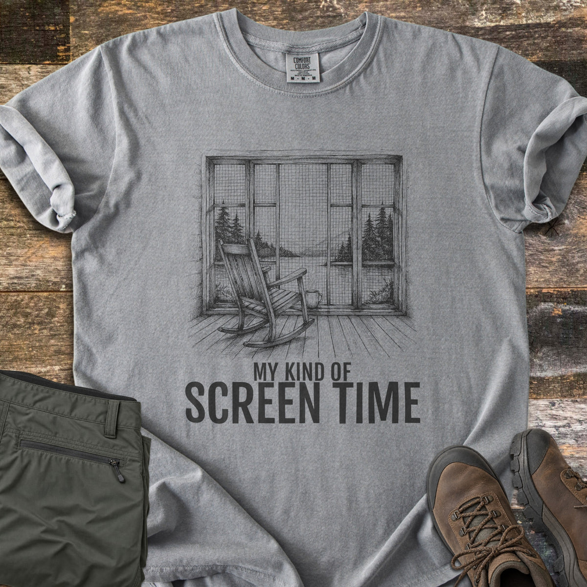 My Kind Of Screen Time Comfort Colors T-shirt in Grey, laid flat. A sketch of a rocking chair on a wooden porch facing a screened window with a lake, pines and distant hills, a mug at its side, above the words My Kind Of Screen Time. Olive hiking pants and brown boots lie on rustic wood.