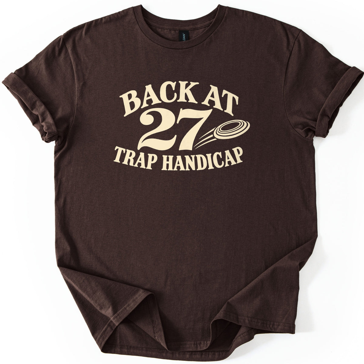 Back at 27 Trap Handicap Comfort Colors T-shirt in Dark Chocolate, laid flat. Cream lettering reads Back at 27 Trap Handicap, with a clay target trailing motion lines beside the large 27. The shirt has rolled sleeves and lies on a white background with the bottom hem tucked under.