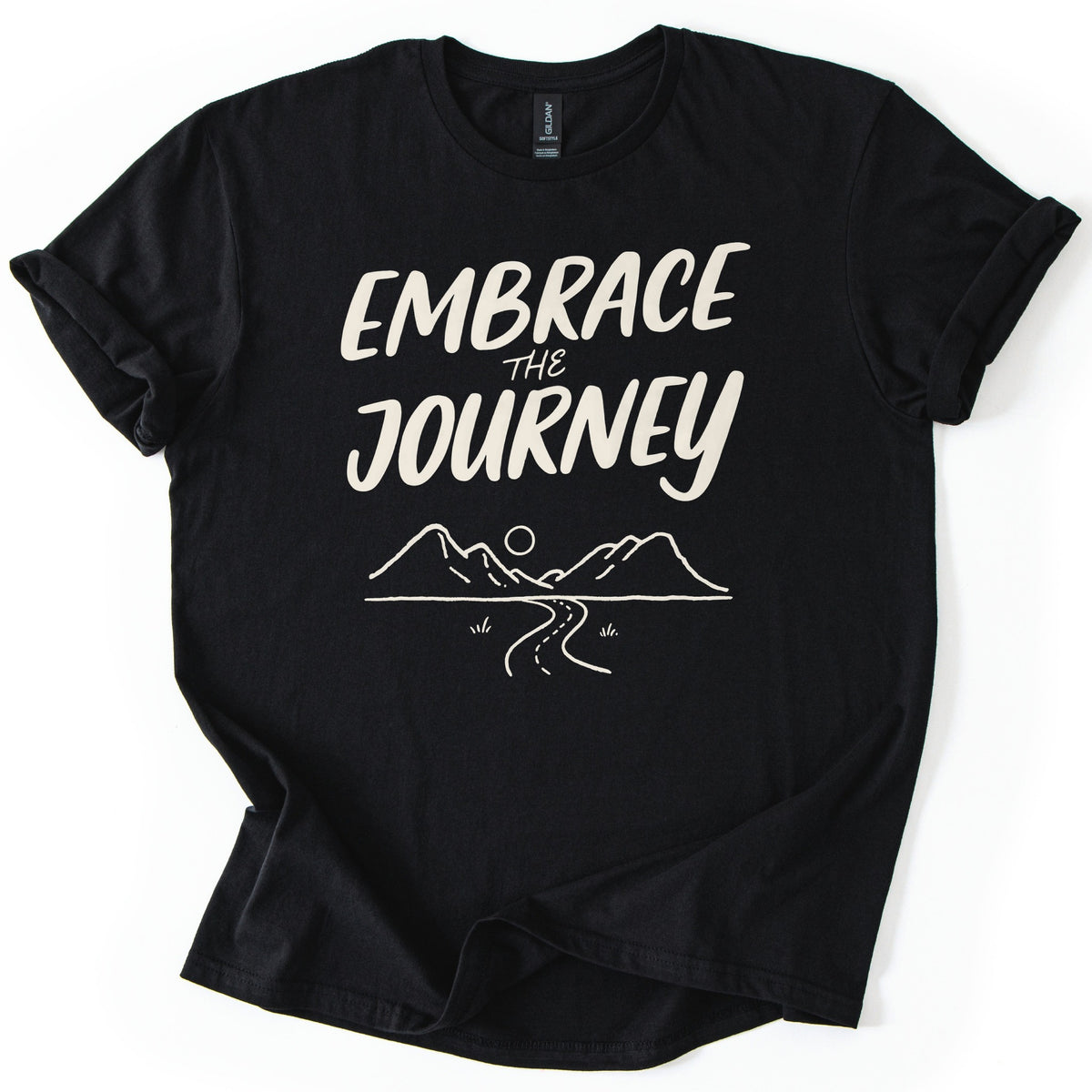 Embrace The Journey Trail Comfort Colors T-shirt in Black, laid flat. Cream hand-lettered words Embrace the Journey sit above a line drawing of two mountains, a round sun, and a winding road with tufts of grass. The sleeves are cuffed, on a white background.