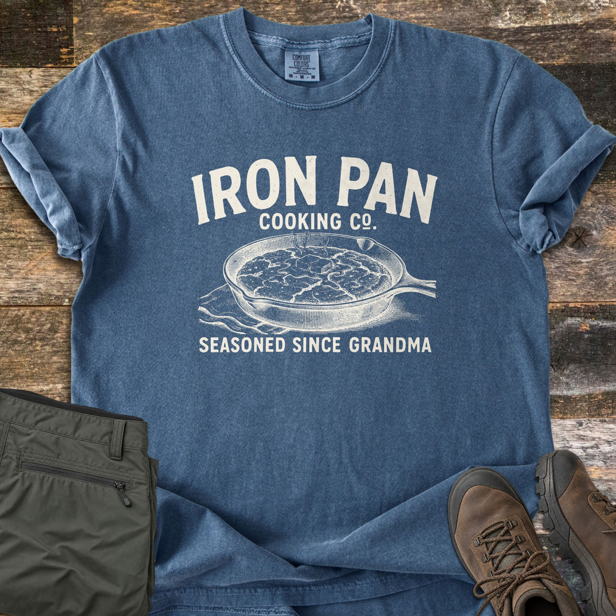 Iron Pan Cooking Co Comfort Colors T-shirt in Denim, laid flat. A vintage-style sketch of a cast iron skillet of cornbread with melting butter, under the words Iron Pan Cooking Co. and above Seasoned Since Grandma. Olive pants and brown hiking boots sit on rustic wood planks.
