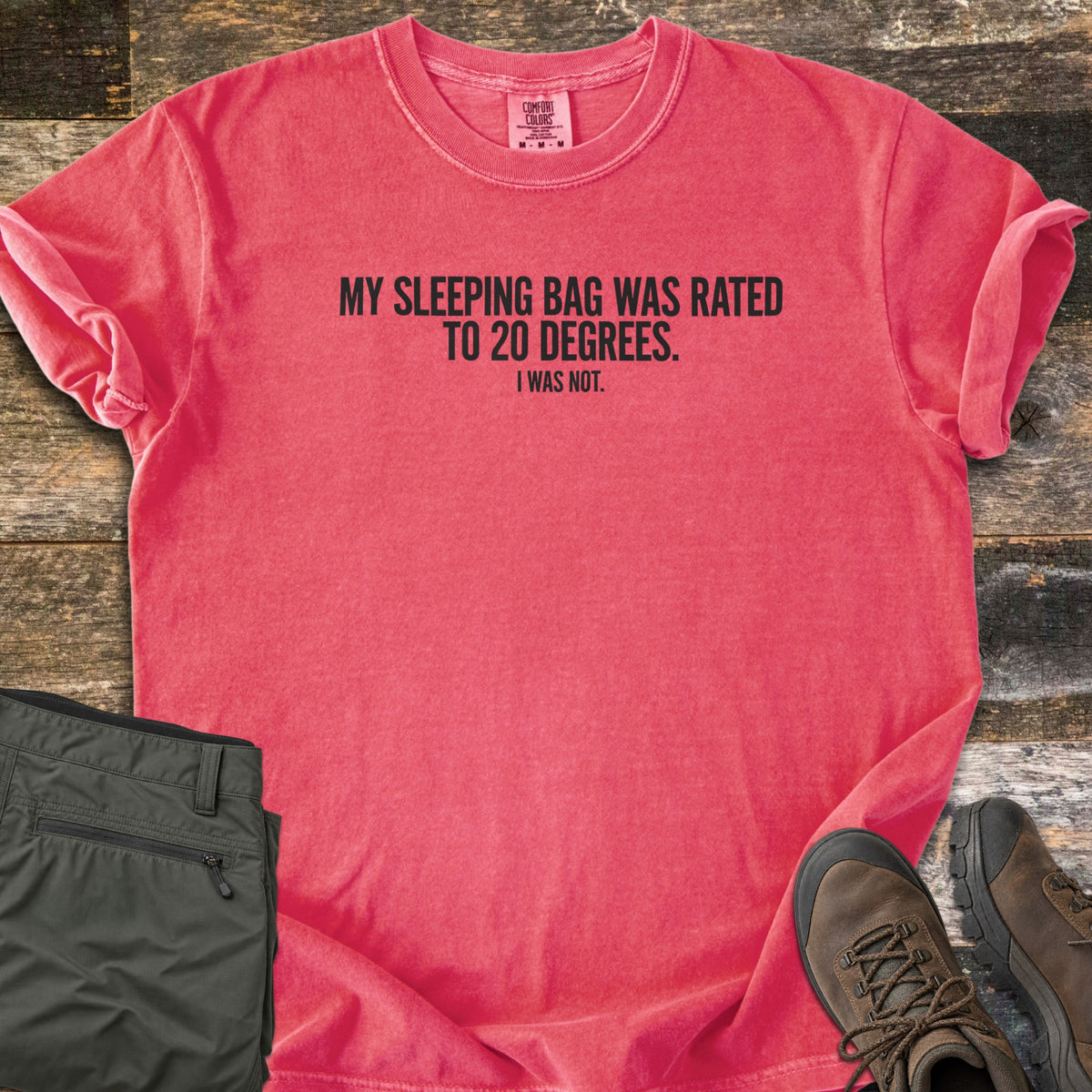 Rated To 20 Degrees Comfort Colors T-shirt in Watermelon, laid flat. Black text across the chest reads My Sleeping Bag Was Rated To 20 Degrees. I Was Not. The shirt has rolled sleeves and lies on weathered wood planks beside folded olive hiking pants and brown hiking boots.