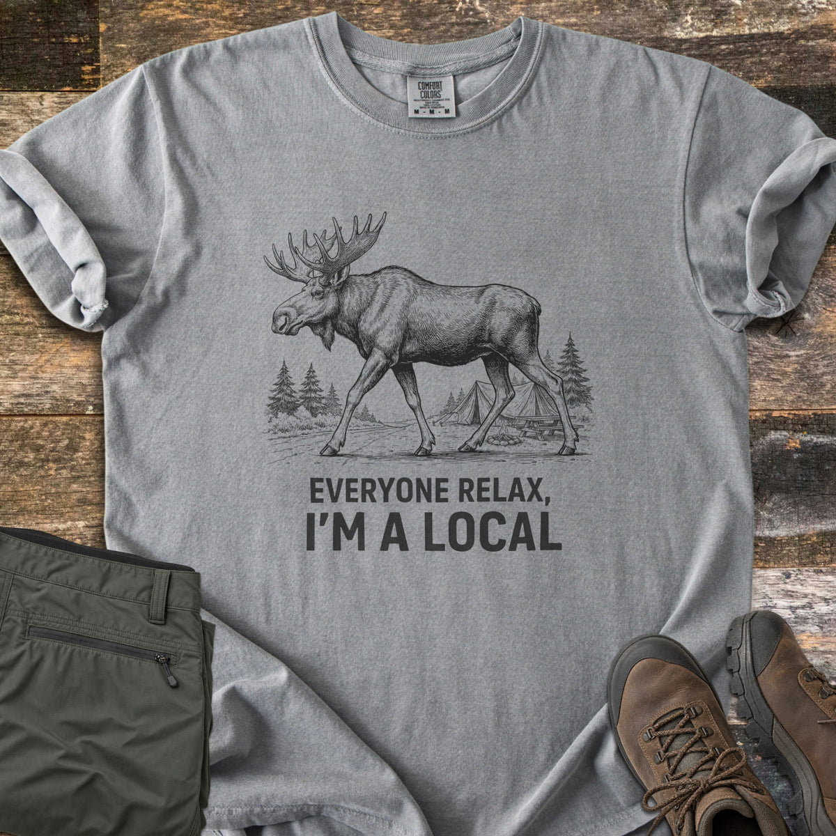 Everyone Relax I'm A Local Comfort Colors T-shirt in Grey, laid flat. A sketched moose walks past pine trees and tents at a campsite above the words Everyone Relax, I'm A Local. Sleeves are rolled on a wood floor beside folded green pants and brown hiking boots.